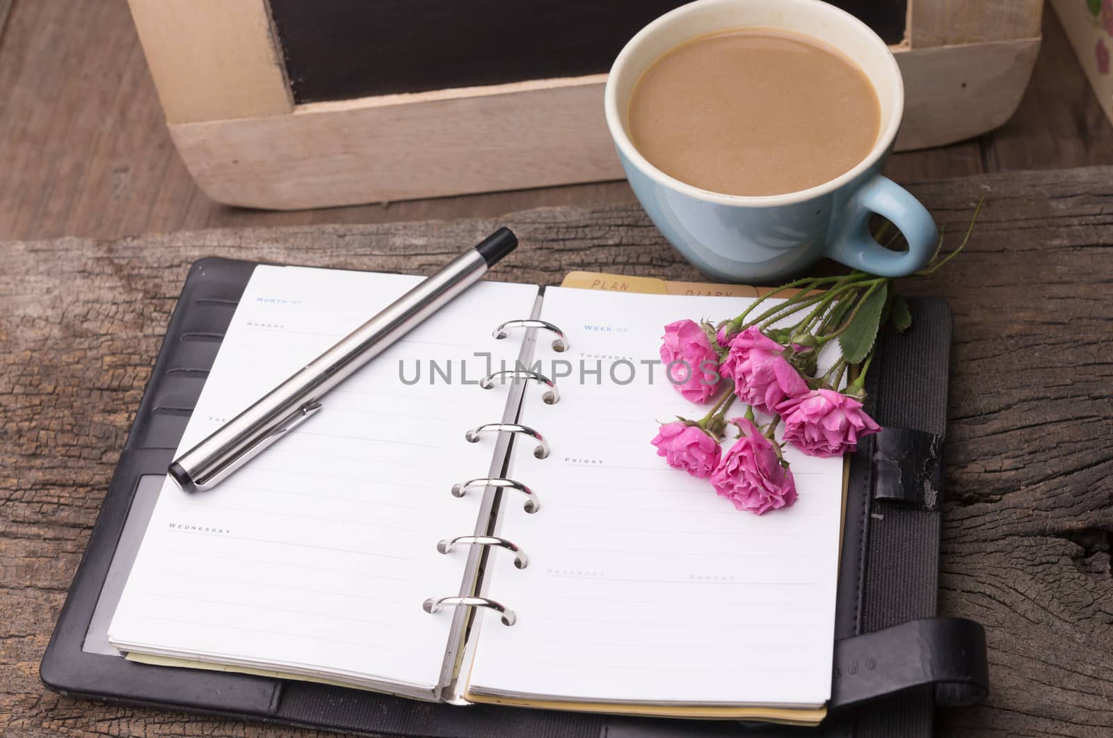 Weekend concept. Pink rose, mug with coffee, diary and pen on a wooden table. Selective focus, copy space background