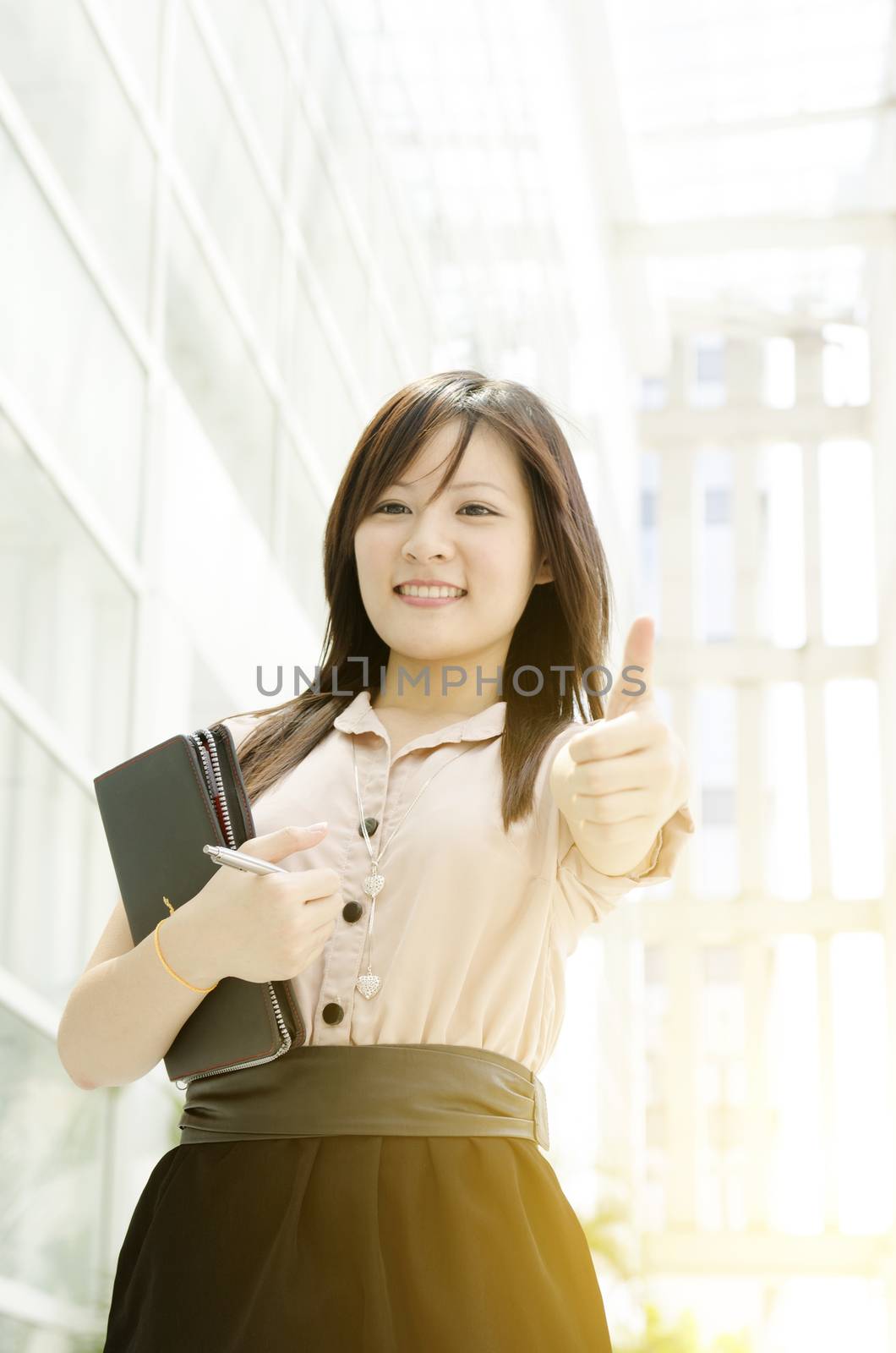 Young Asian business woman smiling and thumb up at an office environment, beautiful golden sunlight at background.