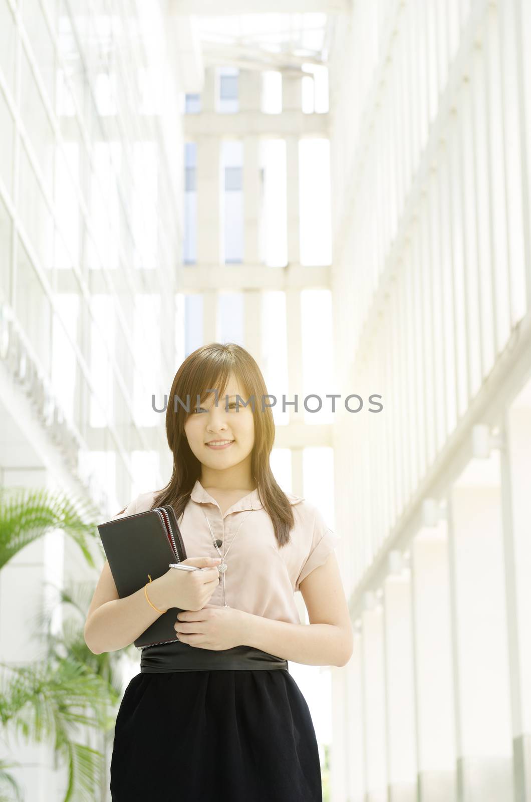 Young Asian business woman smiling and holding diary, standing in an office environment, beautiful golden sunlight at background.