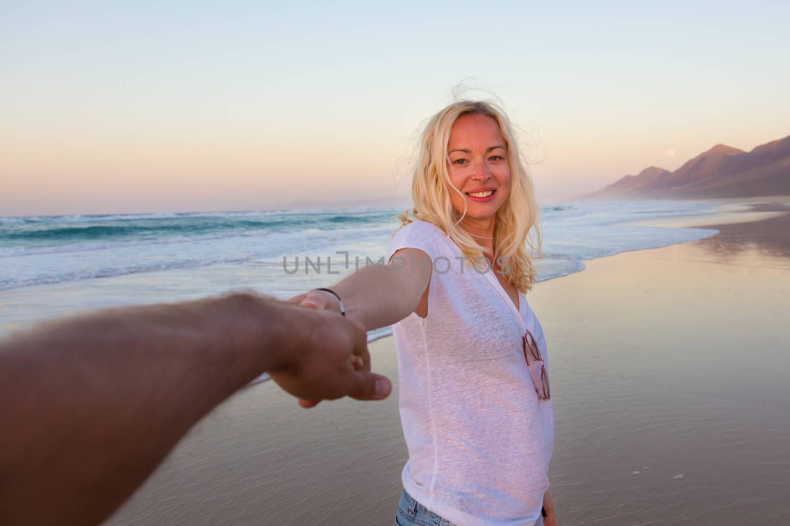 Young romantic couple, holding hands, having fun on perfect deserted beach at sunset. Shot from boyfrieds perspective. Guy looking at her beautiful carefree girlfriend.