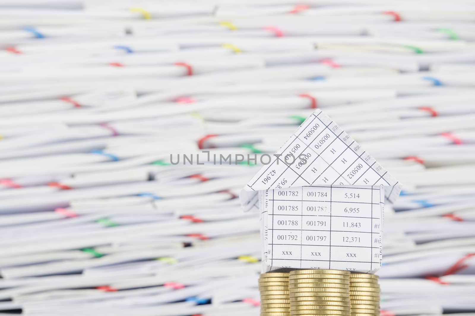 House on pile of gold coins have blur overload of confused paperwork with colorful paperclip as background.