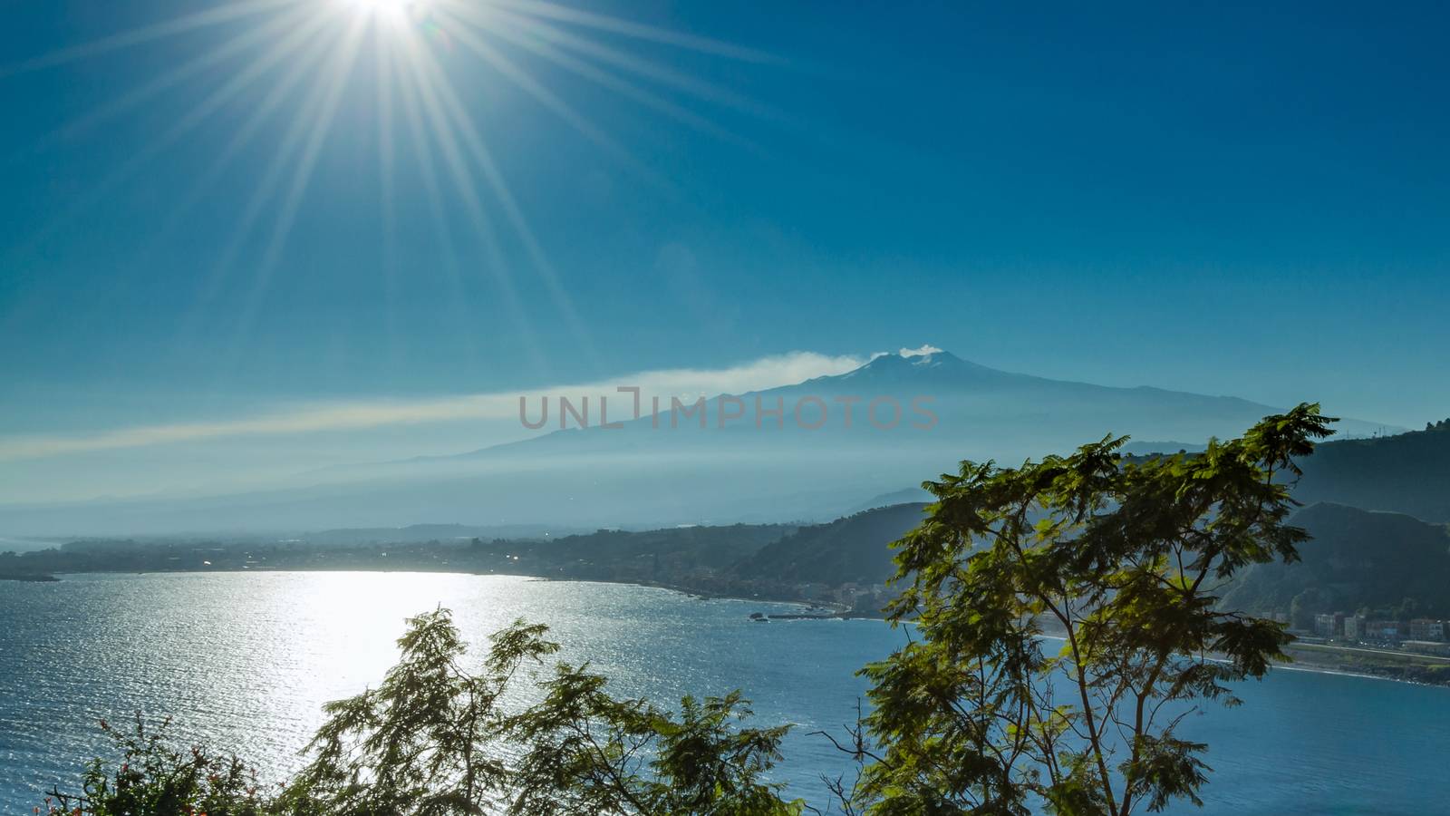 Sicilian seascape from Taormina: mount Etna beyond the blue sea and trees frame on foreground.