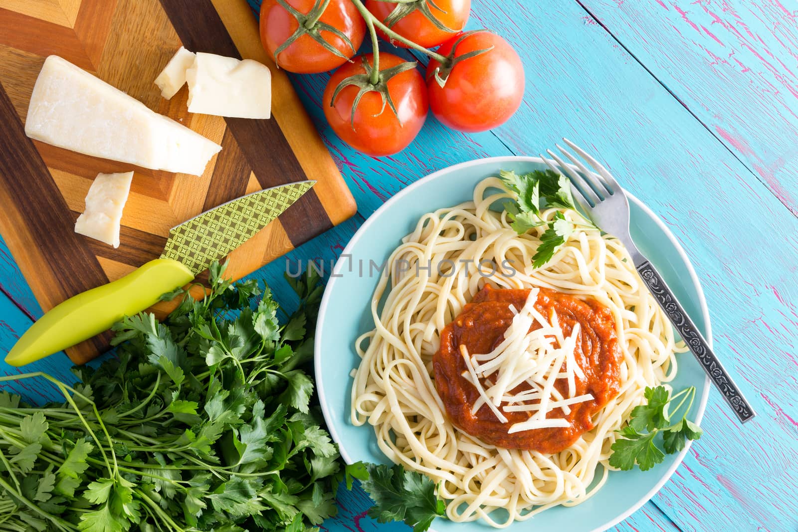 View from above on single plate of freshly prepared spaghetti topped with red sauce, parsley and gruyere cheese next to knife on cutting board with cherry tomatoes over blue table