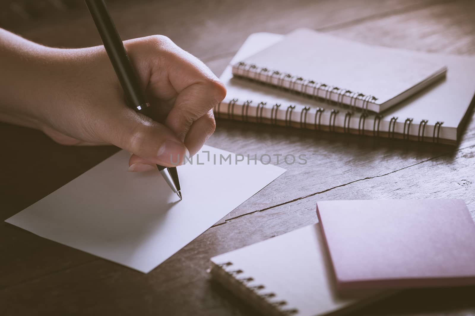 Woman left hand writing on blank paper on wood table with notebooks on wood table with low key scene and vintage filter effect