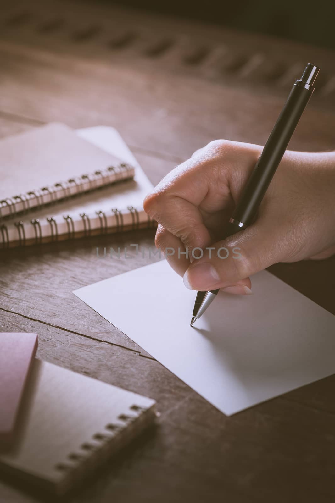 Woman right hand writing on blank paper on wood table with notebooks on wood table with low key scene and vintage filter effect