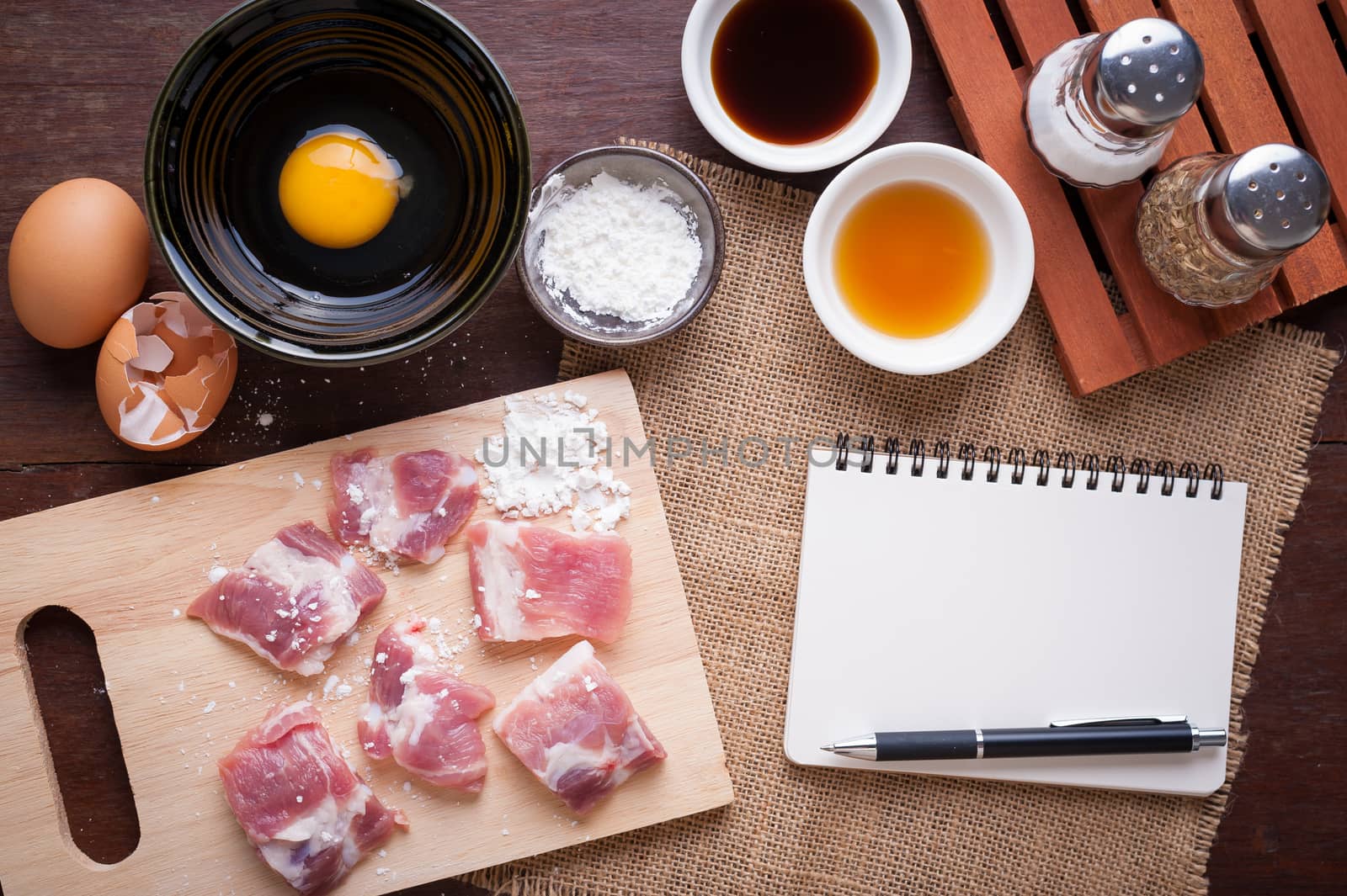 Preparation for cooking in home kitchen. Soft pork ribs on chopping board, notepad for check list, egg, and ingredients on wood table.