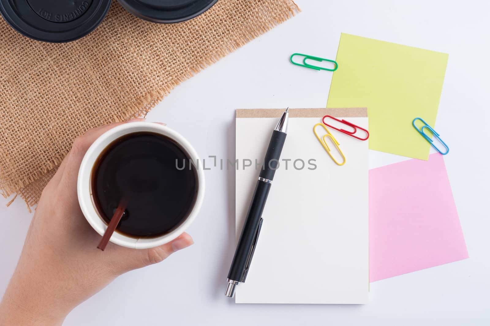 Woman hand holding take away coffee cup beside notepad on office table