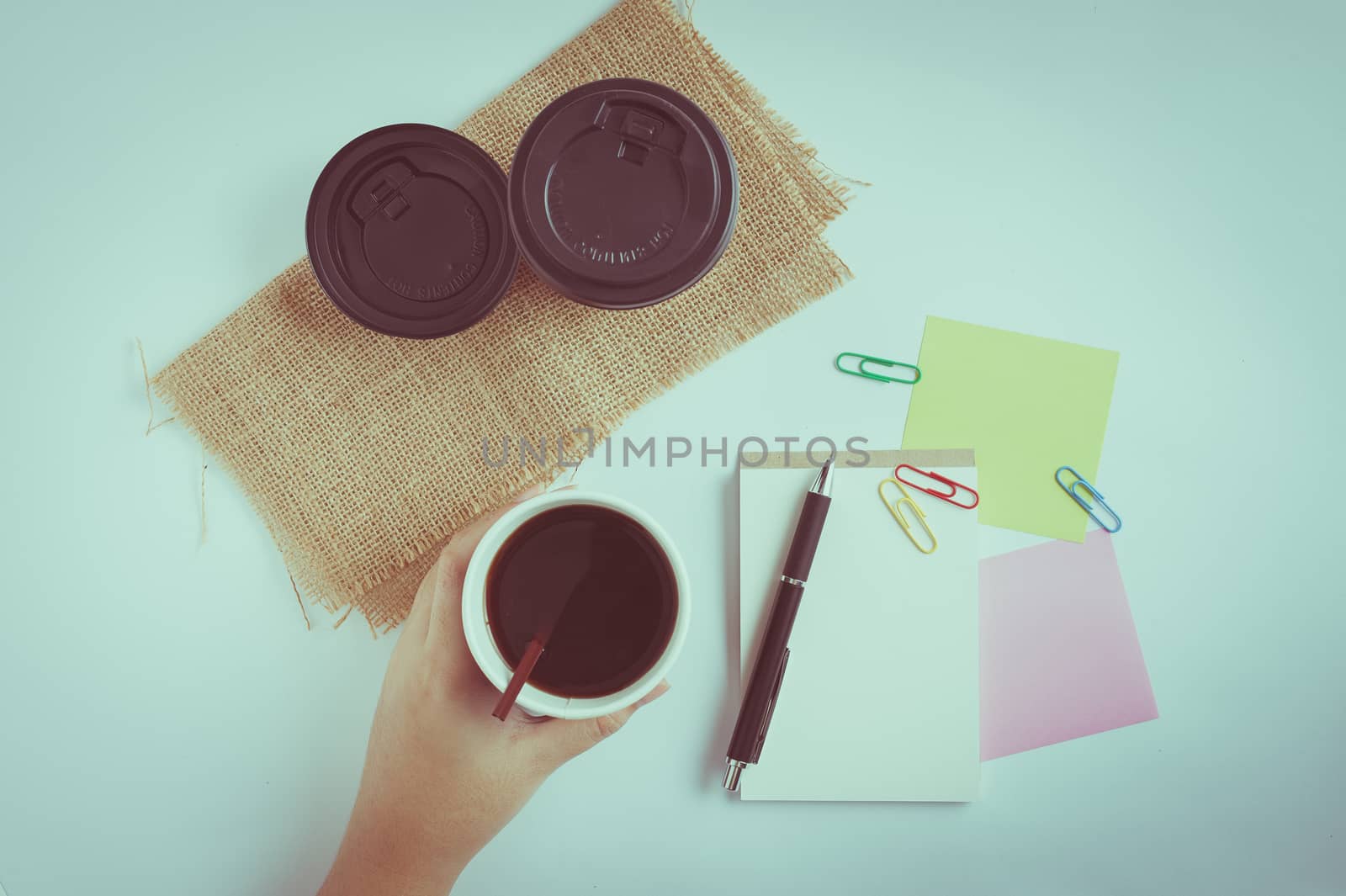 Woman left hand holding take away coffee cup beside notepad on office table with vintage filter effect