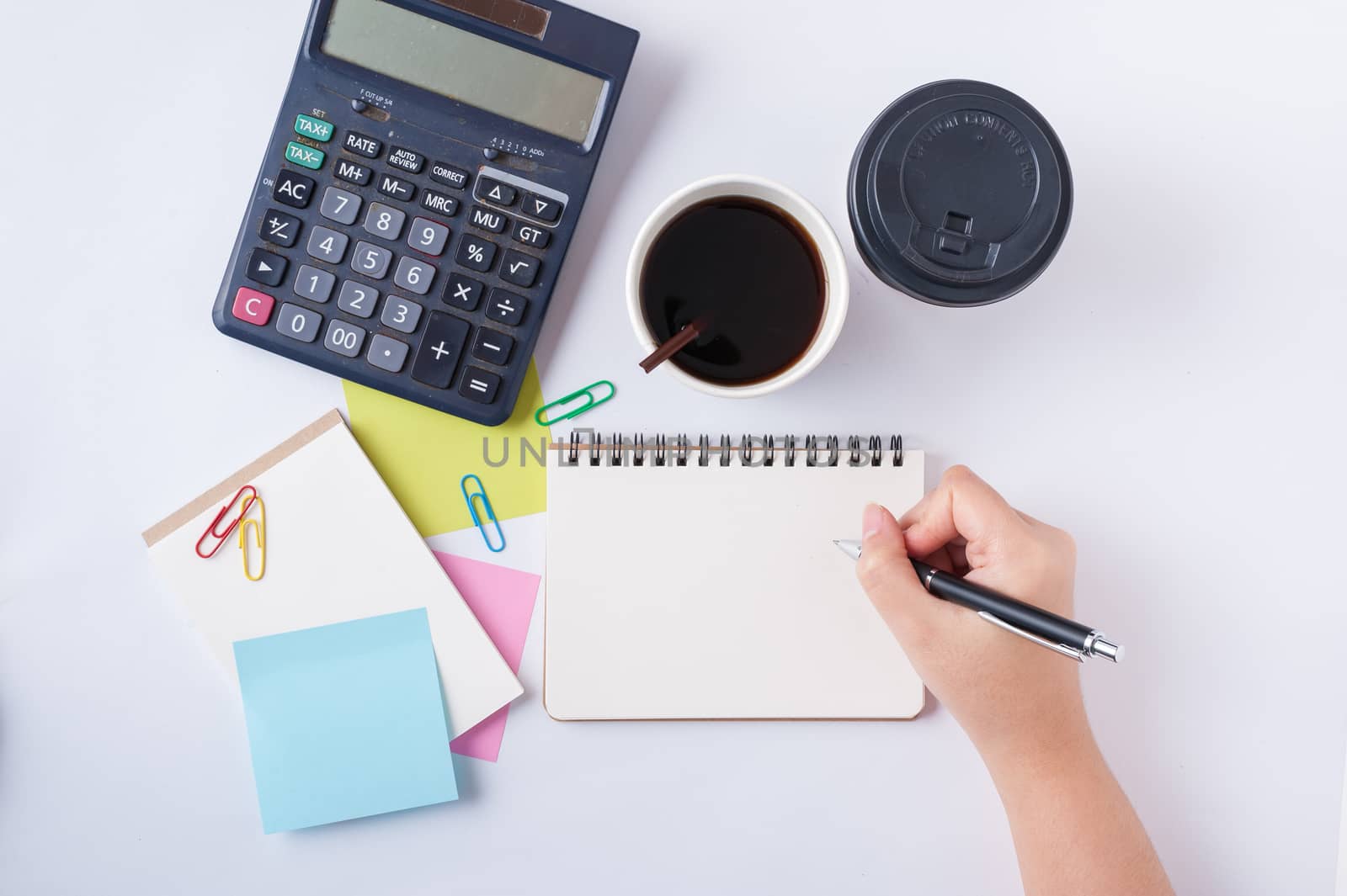 Woman right hand writing on small notepad with blank space for text or message beside sticky paper, calculator, and take away coffee cup on office table