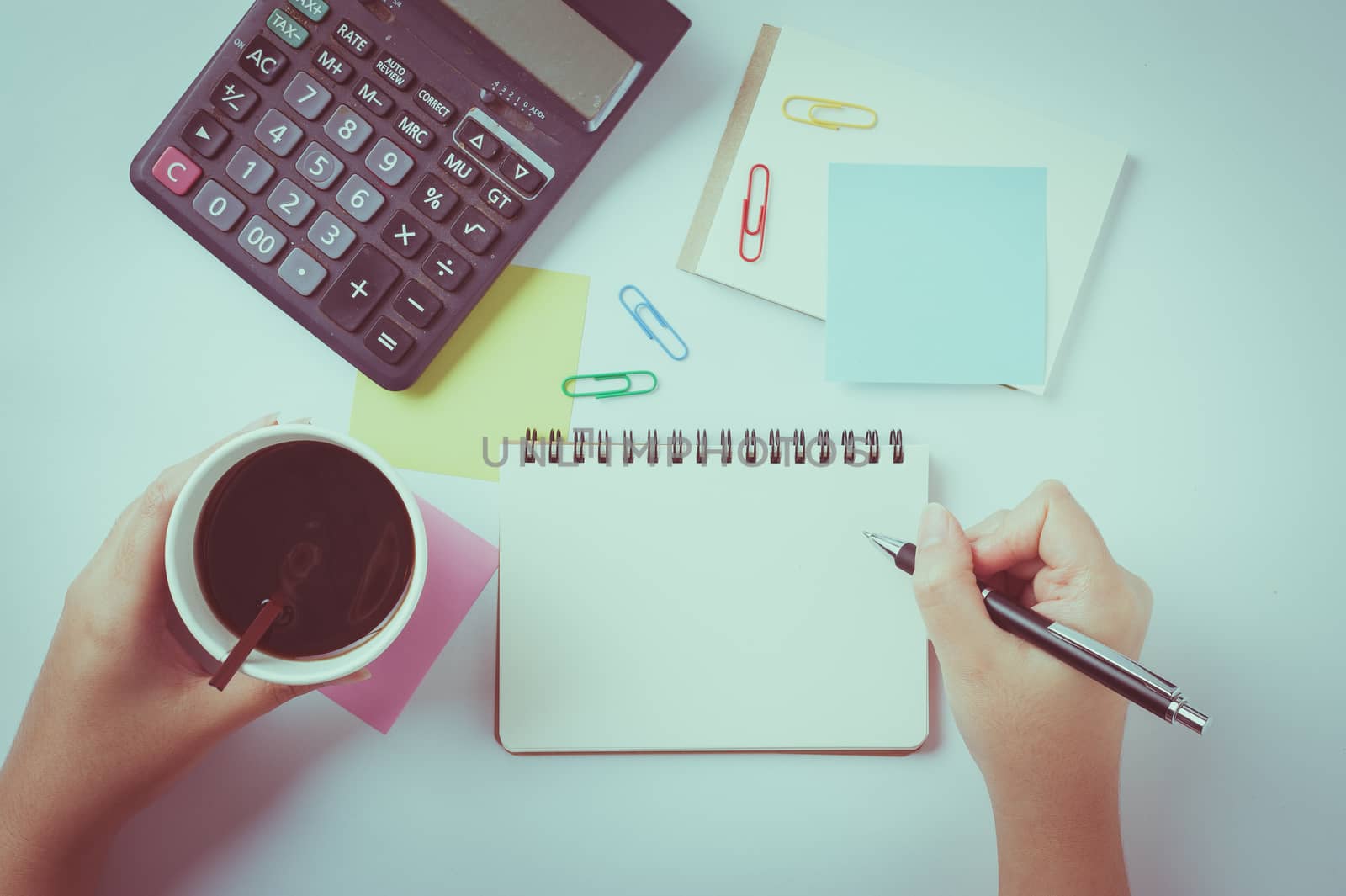Woman right hand writing on small notepad with blank space for text or message beside sticky paper, calculator, and take away coffee cup on office table while another hand holding coffee cup with vintage filter effect
