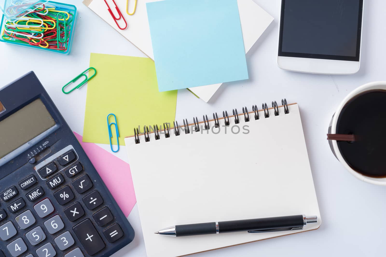 Opened notebook with blank space for text or message, sticky paper, calculator, smart phone, and take away coffee cup on office table