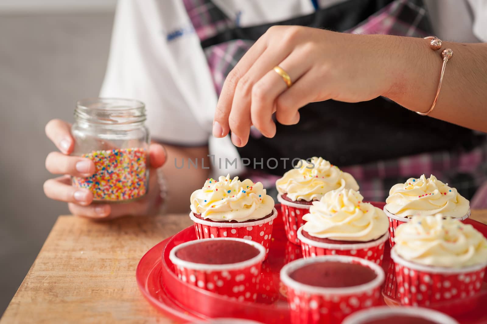The action of making red velvet cupcakes. Woman hand adding sugar sprinkle on cupcake.