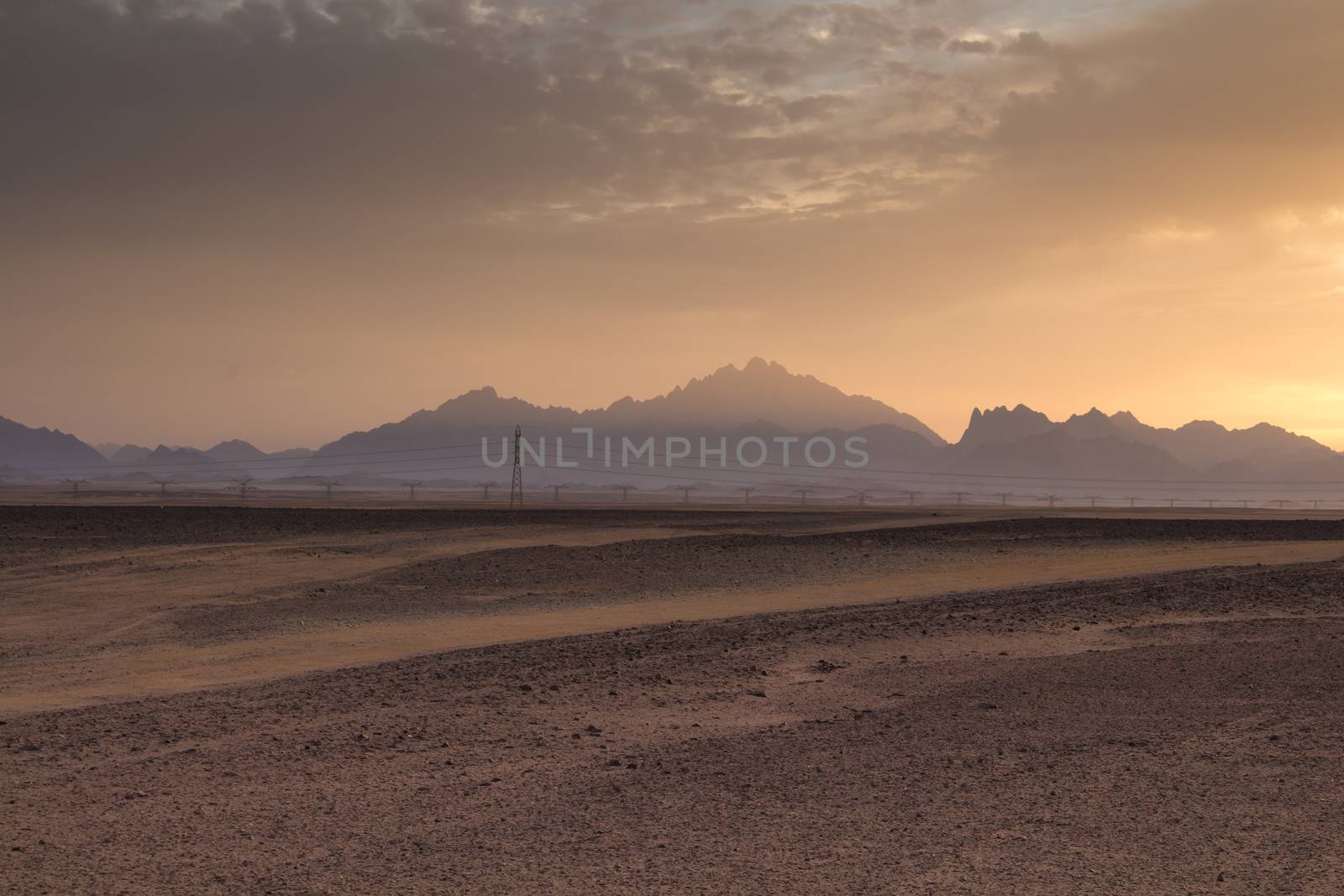 Sandy land in the foreground, rocky mountains in the background. Little clouds on the sky during the sunset.