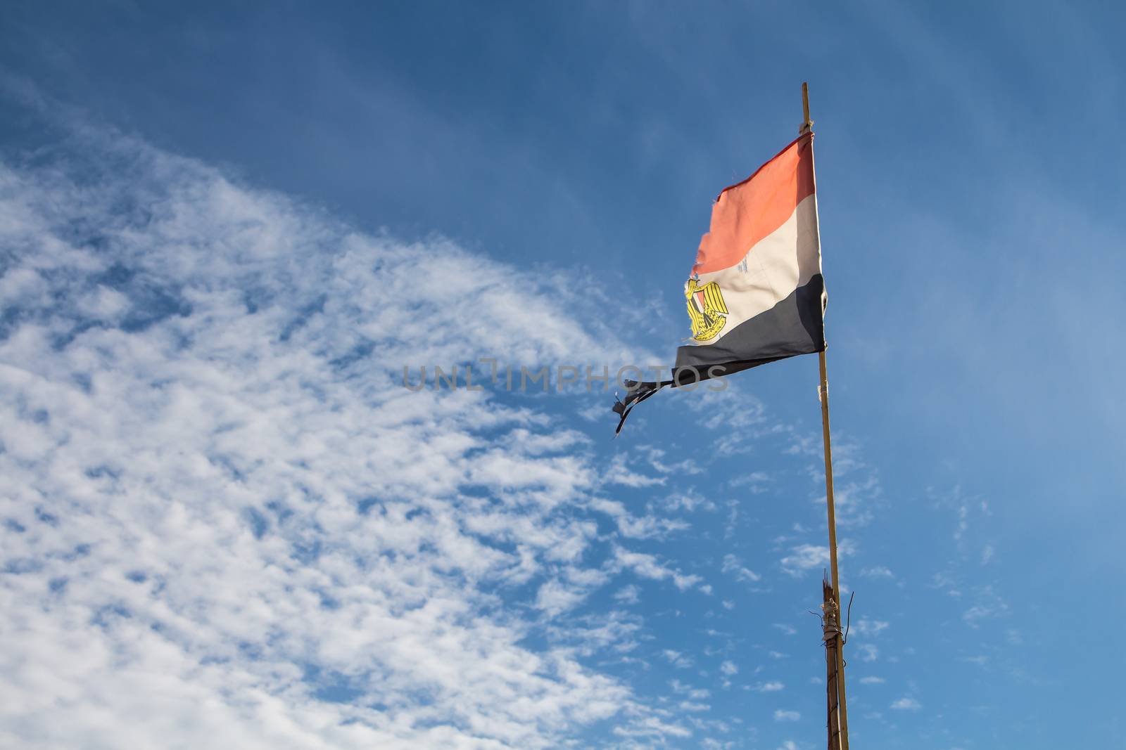 Ragged egyptian flag in the wind, enlightened by sunlight. Blue sky with intense white clouds on the left side.