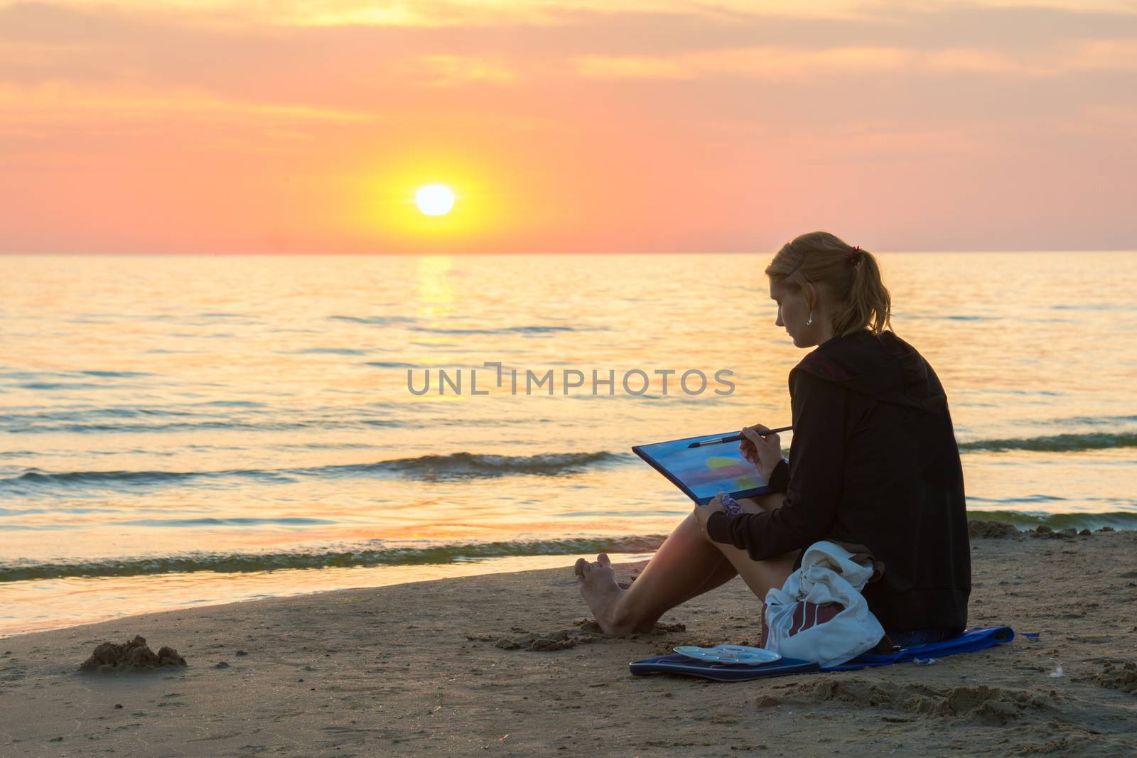 Young girl sitting on the beach and water paints watercolor landscape with sunset on a piece of paper
