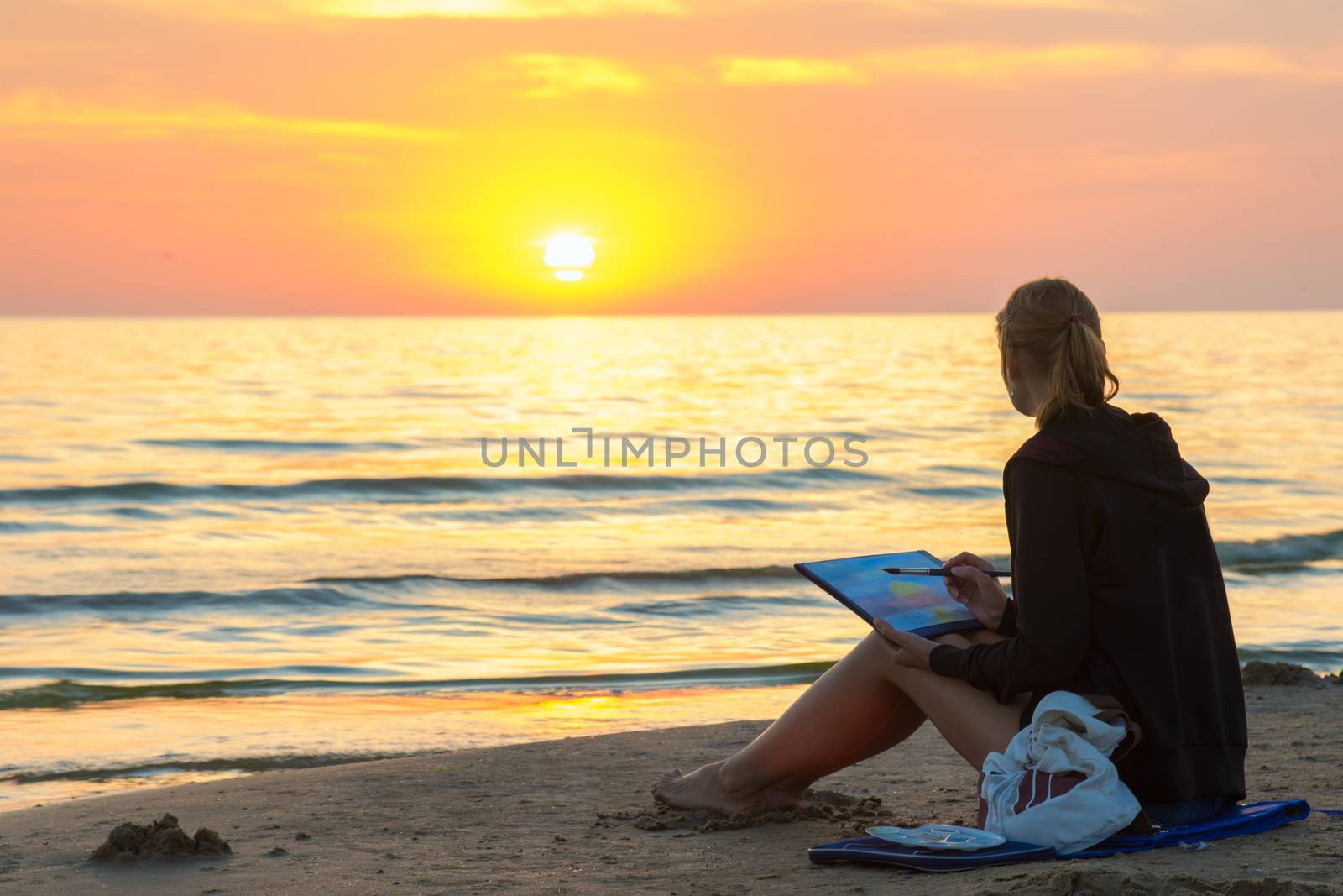 Young girl sitting on the beach and water paints watercolor landscape with sunset on a piece of paper
