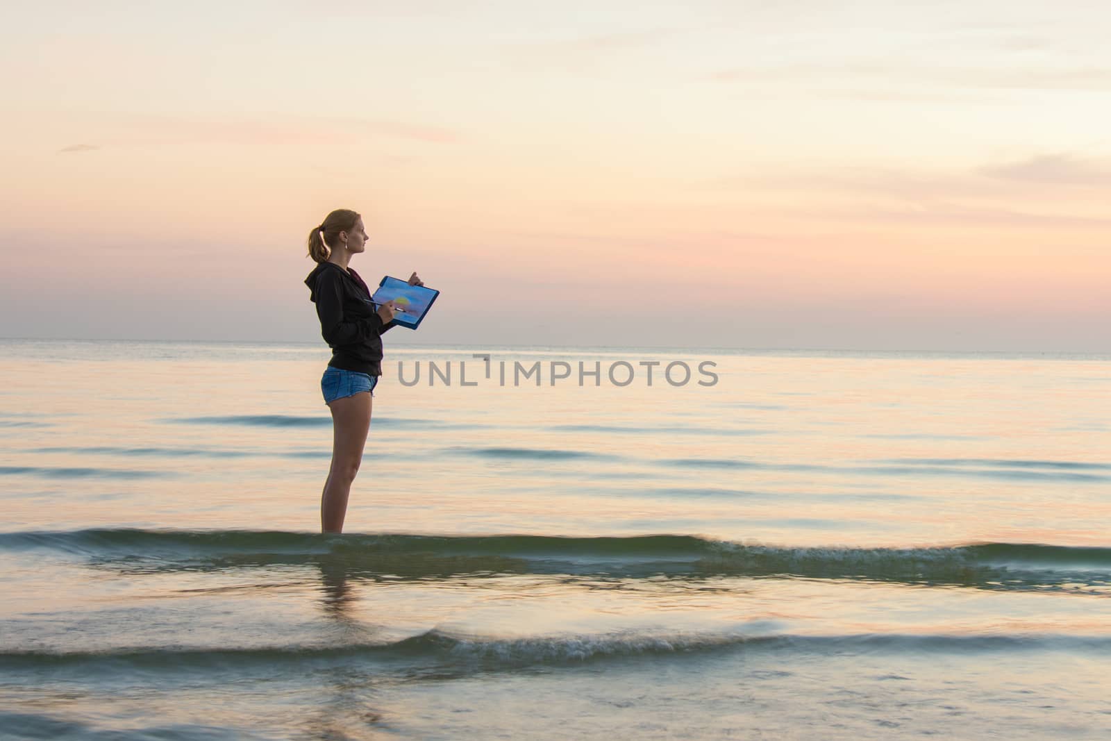 Young girl standing in the water on the beach, and draws on the colors of the landscape with sunset on a piece of paper