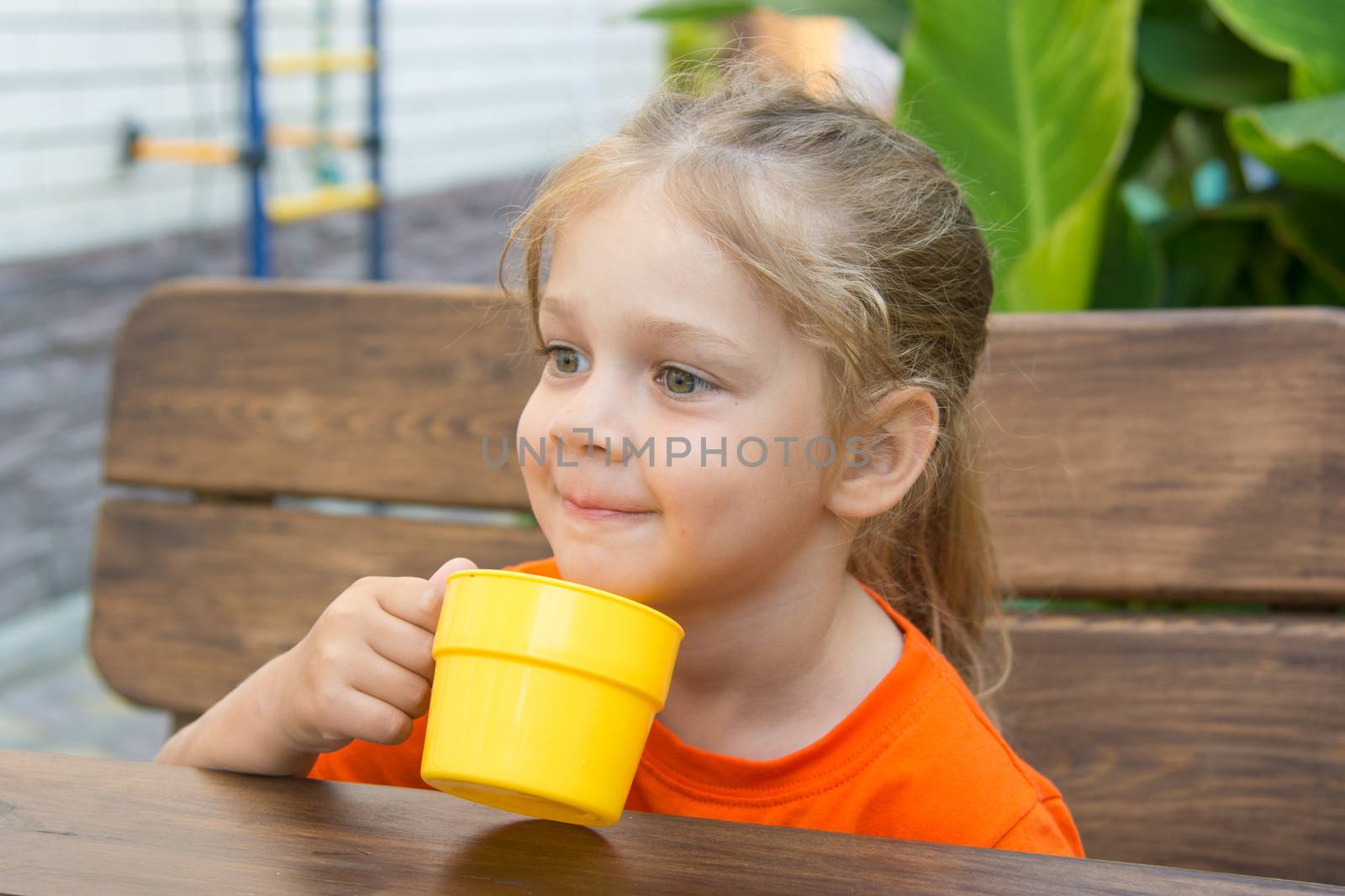 Four-year girl sitting at a wooden table on the porch and drink from the cup