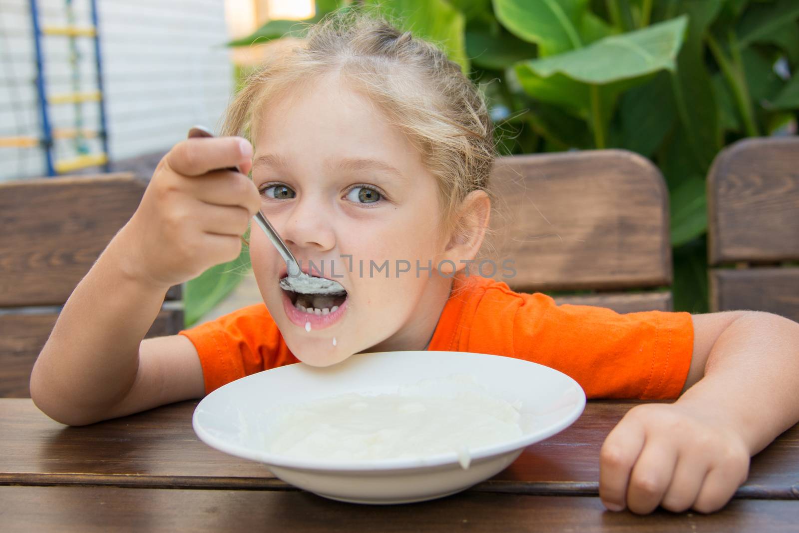Five-year girl sits at a table on the veranda and enjoy eating porridge