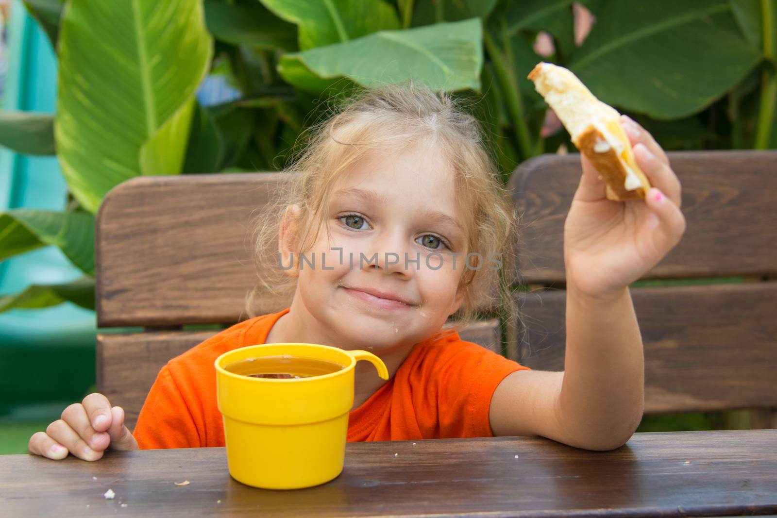 Four-year girl sitting at a wooden table on the porch, drinking a drink from the cup and eating a bun
