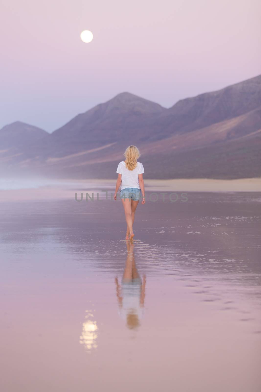 Woman walking on sandy beach in dusk leaving footprints in the sand. Beach, travel, concept. Copy space. Vertical composition.