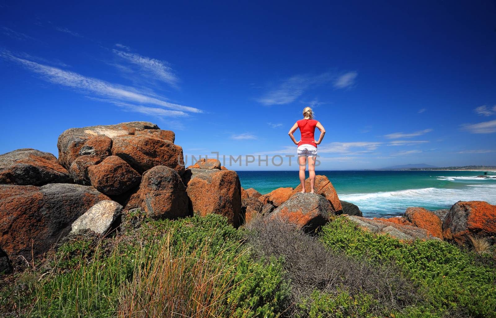 A female standing on the vibrant orange rocks and looking out to sea.  Bingie Point, Eurobodalla National Park on the Sapphire Coast of NSW, Australia.  Mount Gulaga in the far distance
