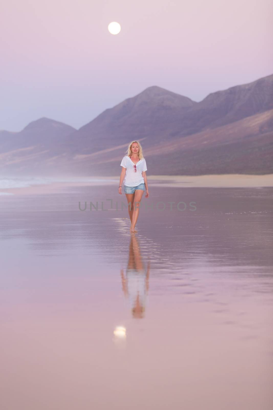 Woman walking on sandy beach in dusk leaving footprints in the sand. Beach, travel, concept. Copy space. Vertical composition.