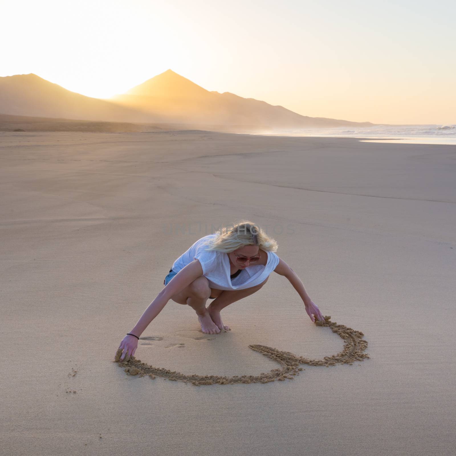 Lady drawing heart shape in sand on beach. by kasto