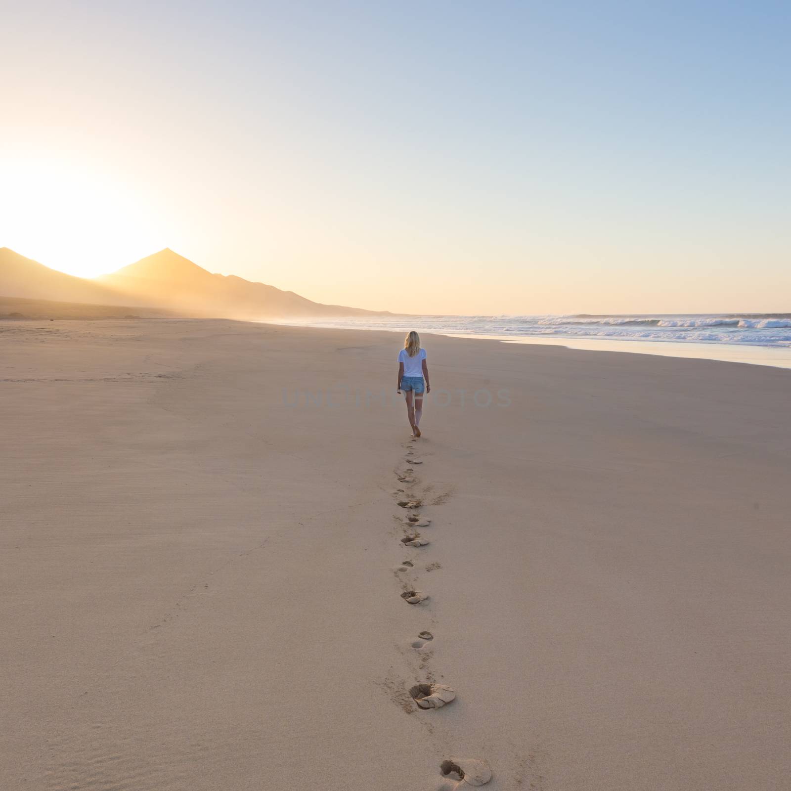 Lady walking on sandy beach in sunset leaving footprints behind. by kasto