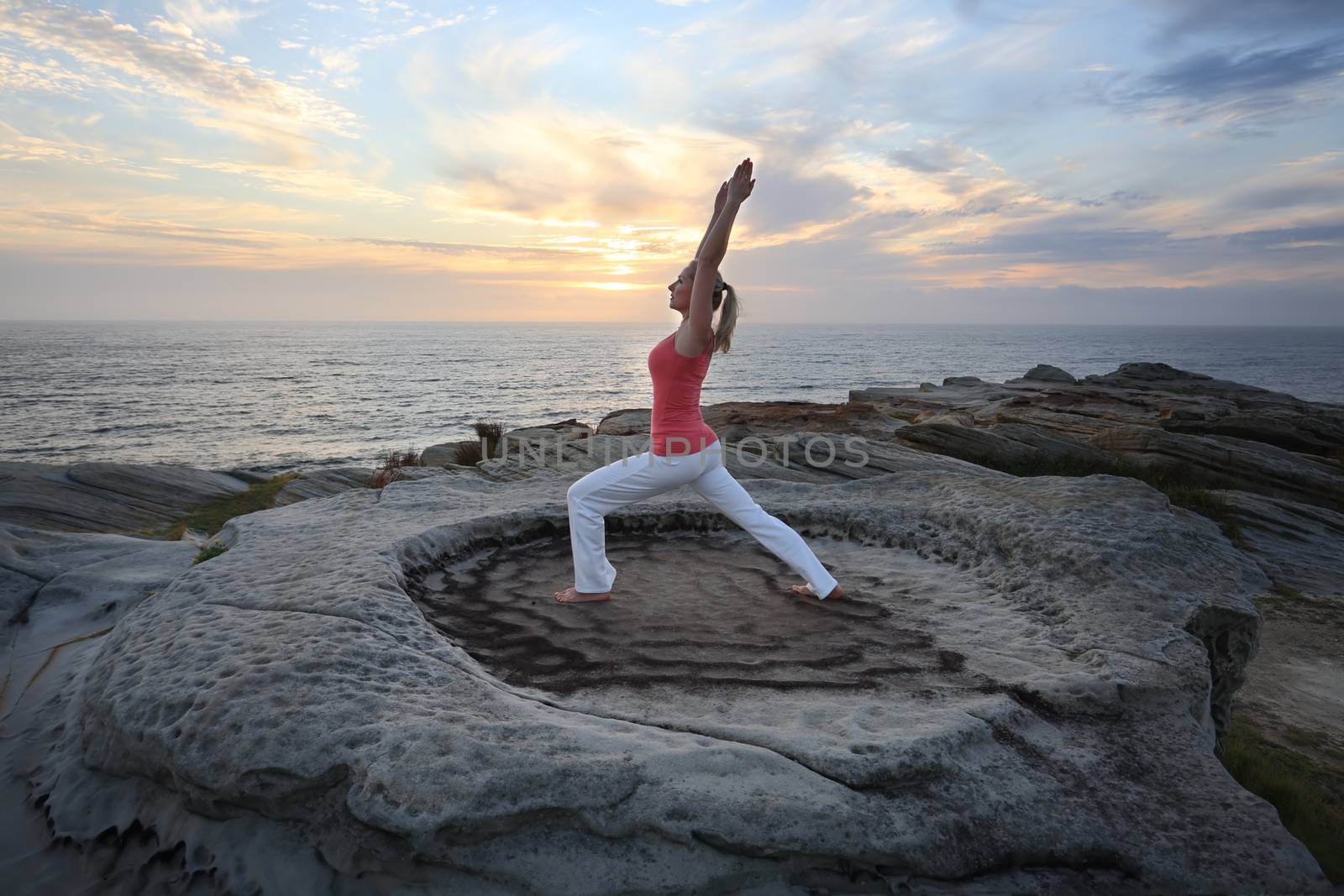 A female lunges forward while stretching the other leg and arms straight above her head as a yoga pilates routine by the sea. A variation of anjaneyasana, ashwa sanchalasana.