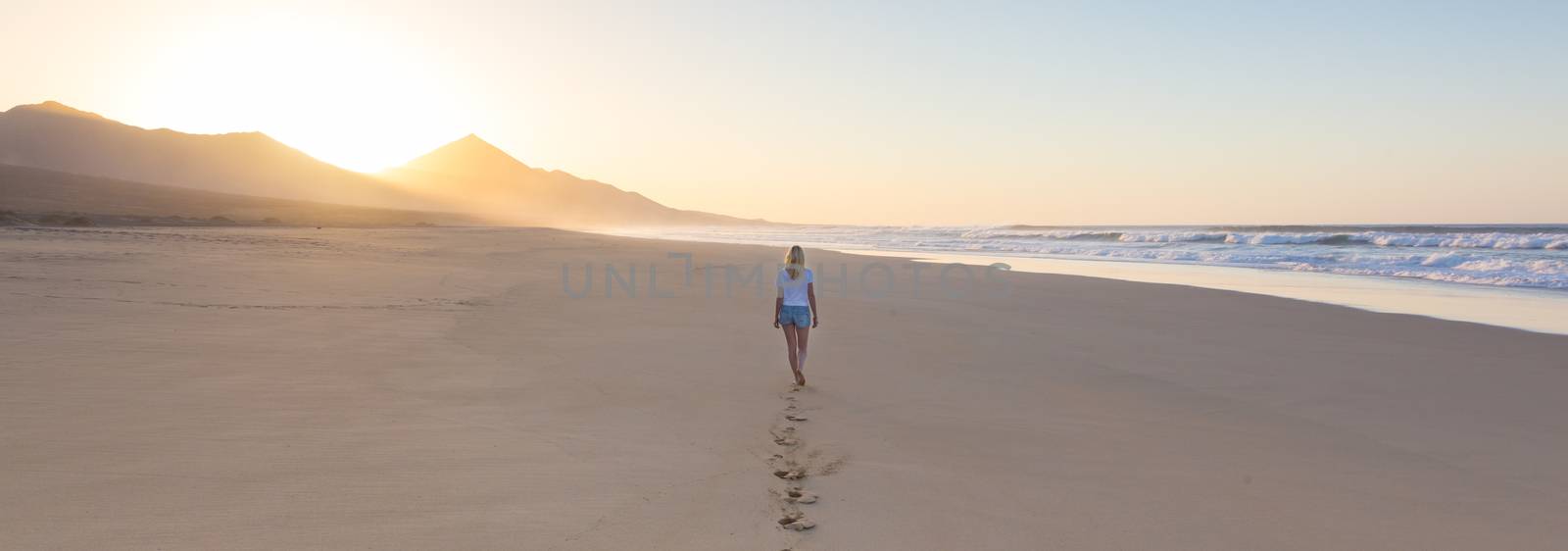 Lady walking on sandy beach in sunset leaving footprints behind. by kasto