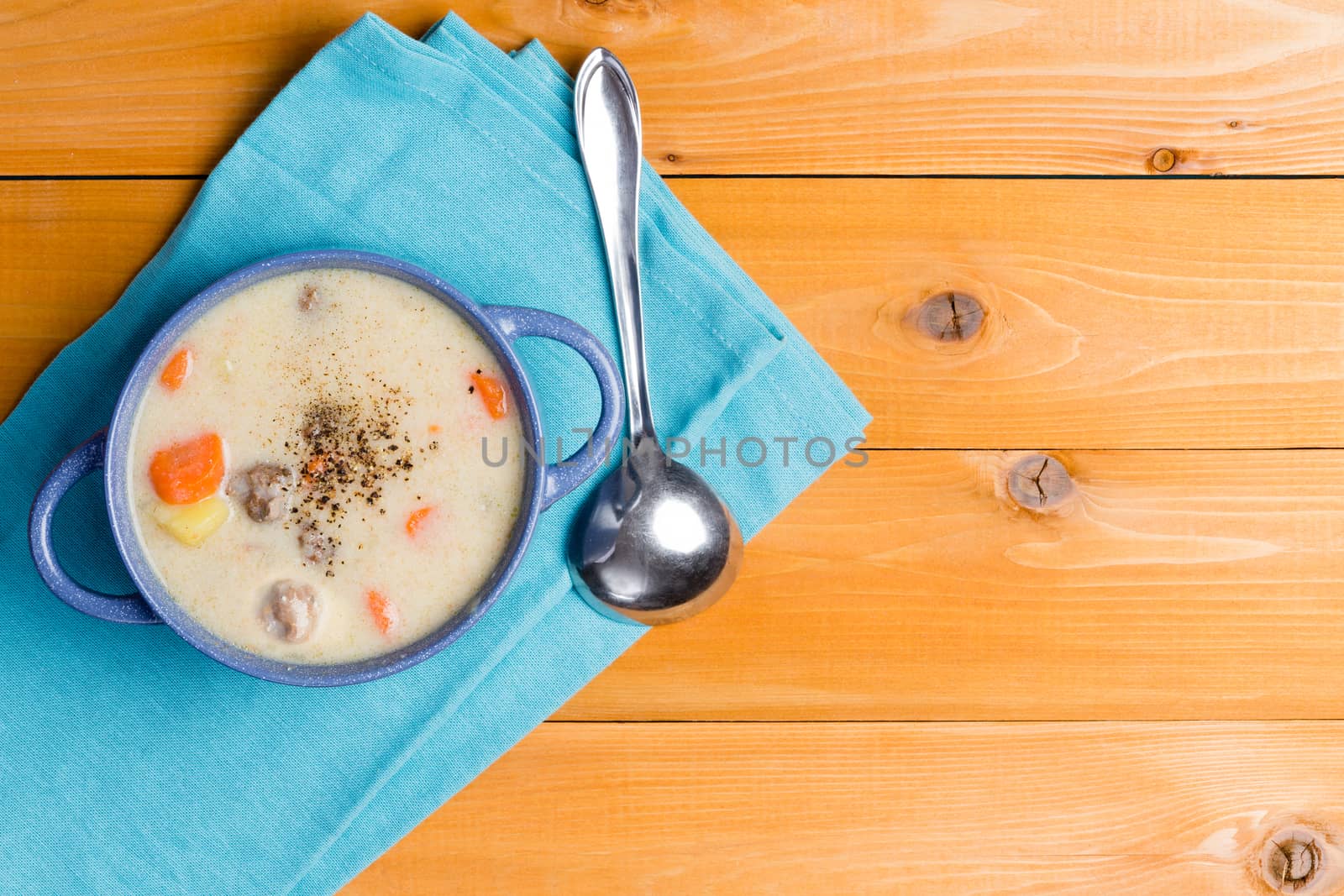 Tasty starter of homemade meatball soup with fresh vegetables seasoned with spicy ground black pepper and served on a blue napkin on a wood table with copy space, overhead view