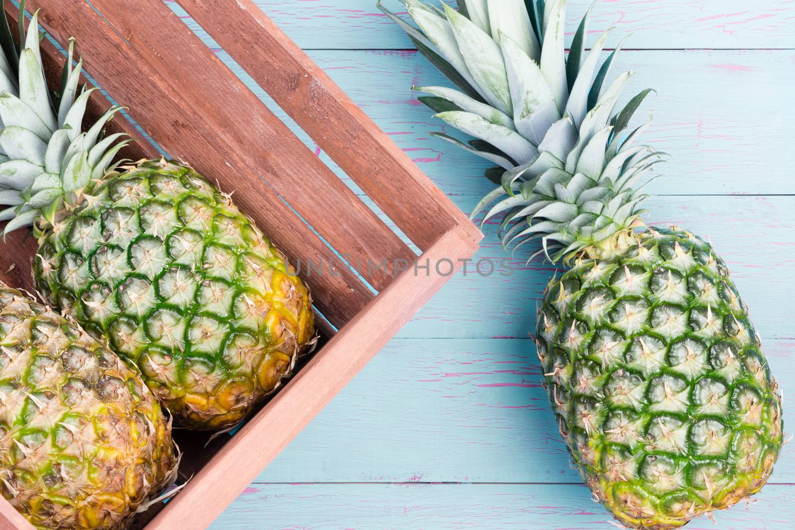 Three fresh pineapples, two in a wooden crate and one alongside on a blue wooden table lying on their sides viewed close up from above
