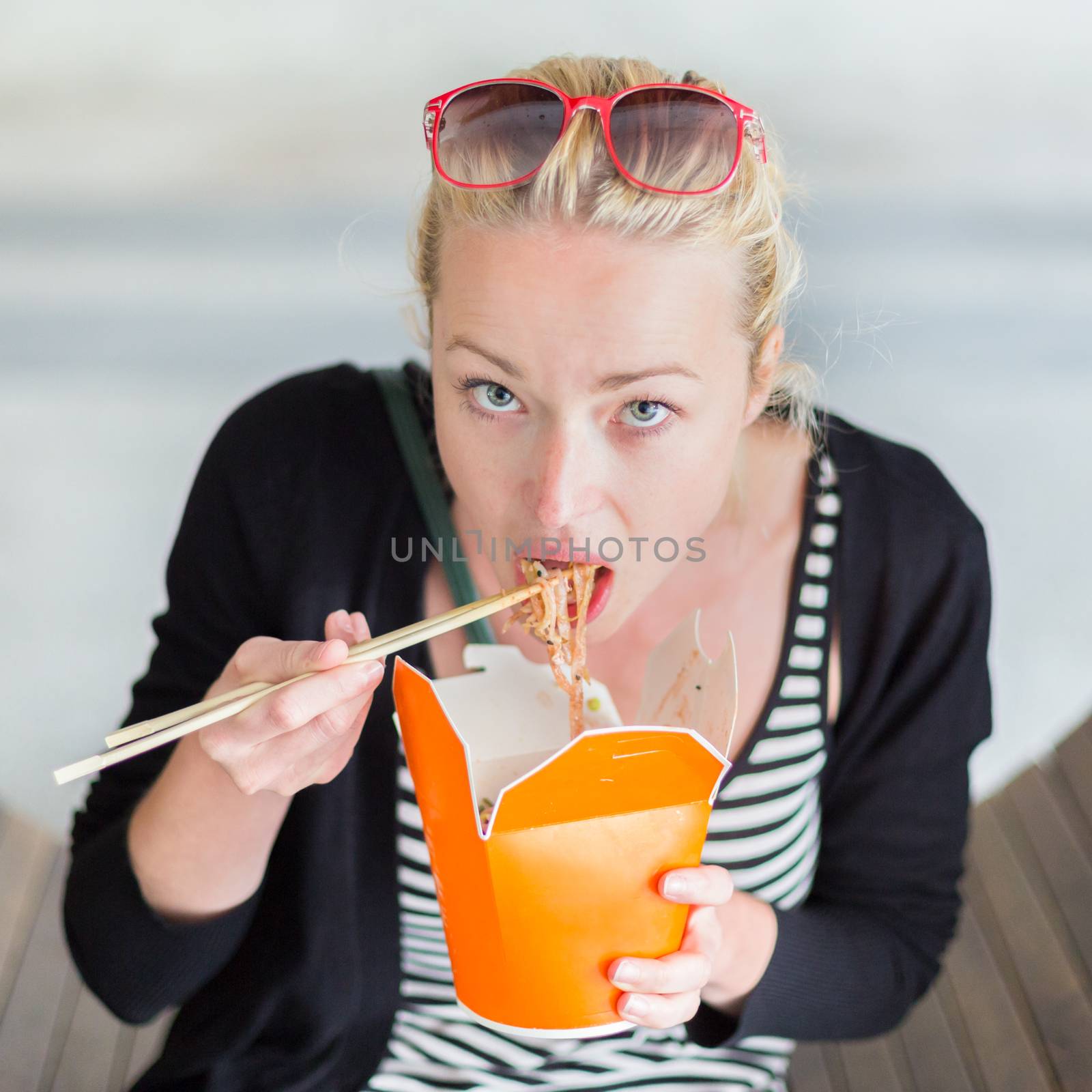 Beautiful young woman, sitting on the bench in park, holding a fast food lunch box, eating noodles from Chinese take-away with traditional wooden chopsticks.