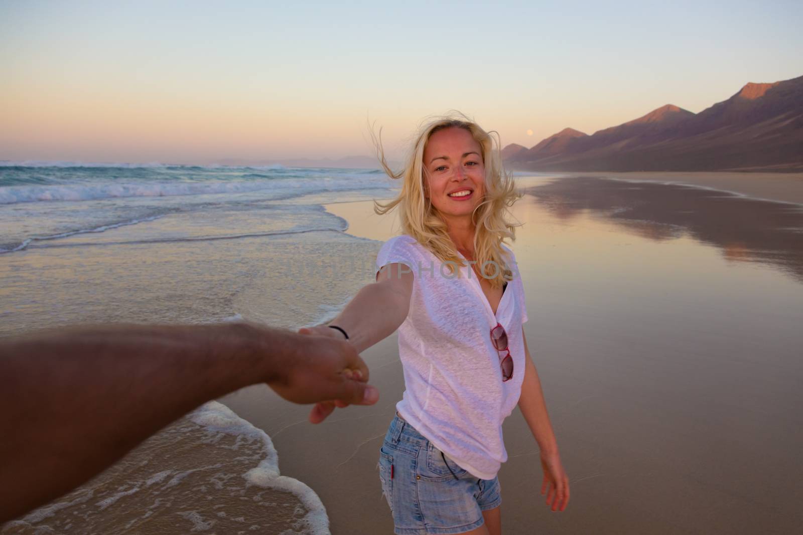 Young romantic couple, holding hands, having fun on perfect deserted beach at sunset. Shot from boyfrieds perspective. Guy looking at her beautiful carefree girlfriend.