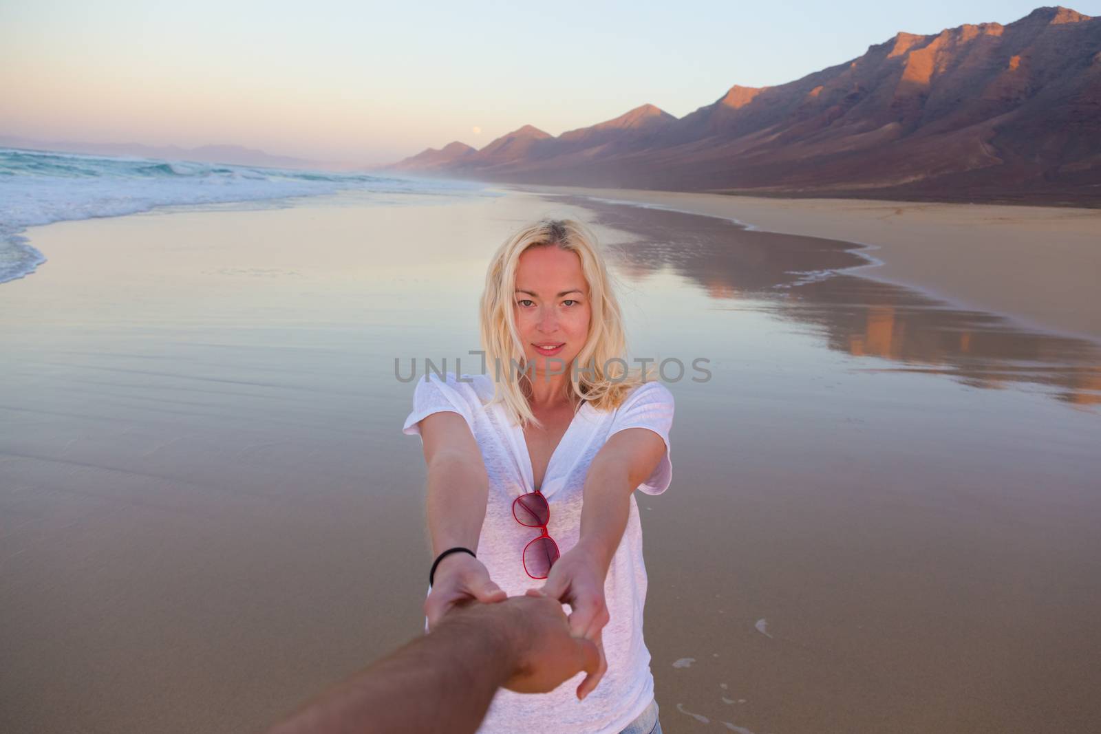Young romantic couple, holding hands, having fun on perfect deserted beach at sunset. Shot from boyfrieds perspective. Guy looking at her beautiful carefree girlfriend.