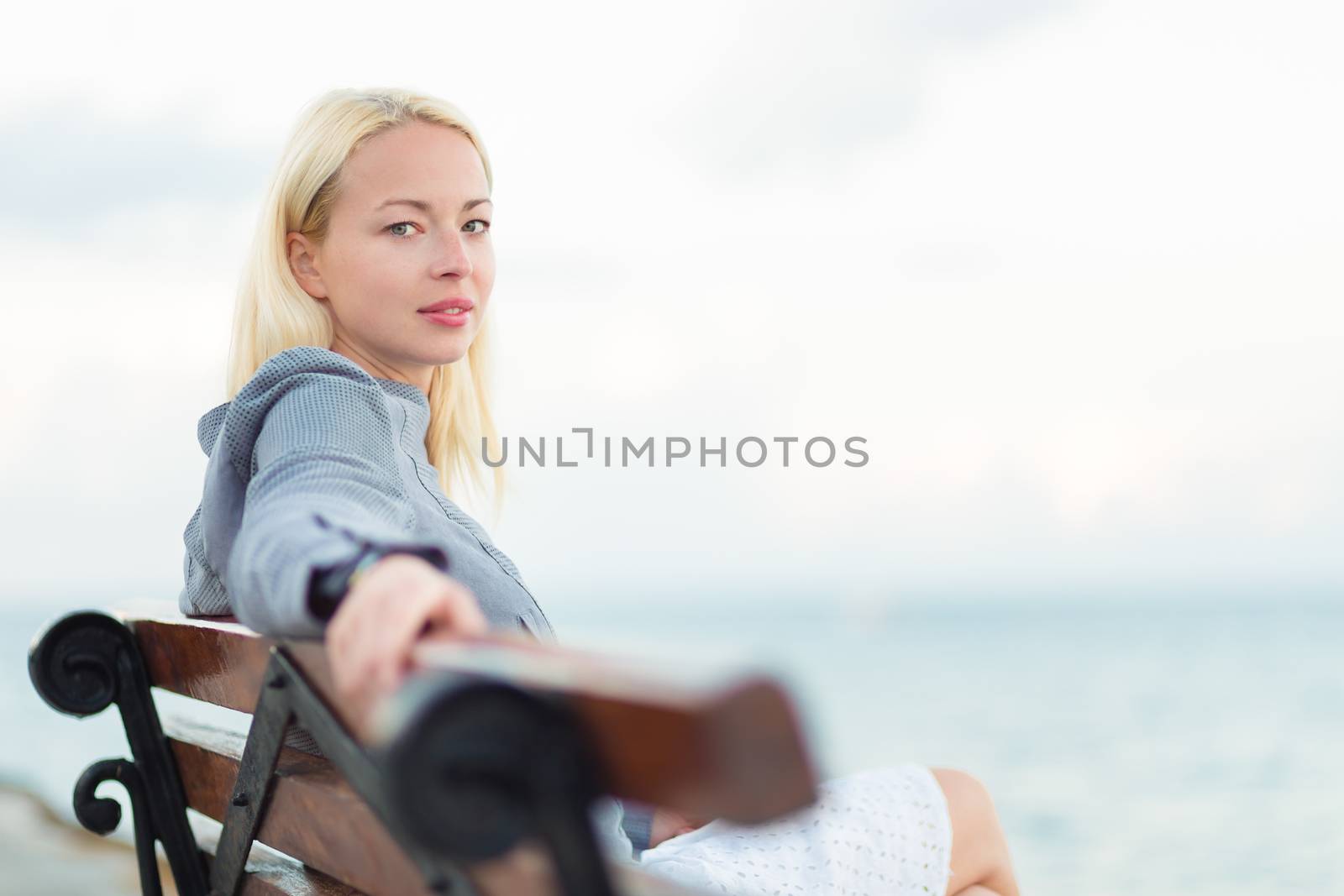 Beautiful woman sitting on a vintage wooden bench, relaxing on fresh breeze by the sea, looking at camera.