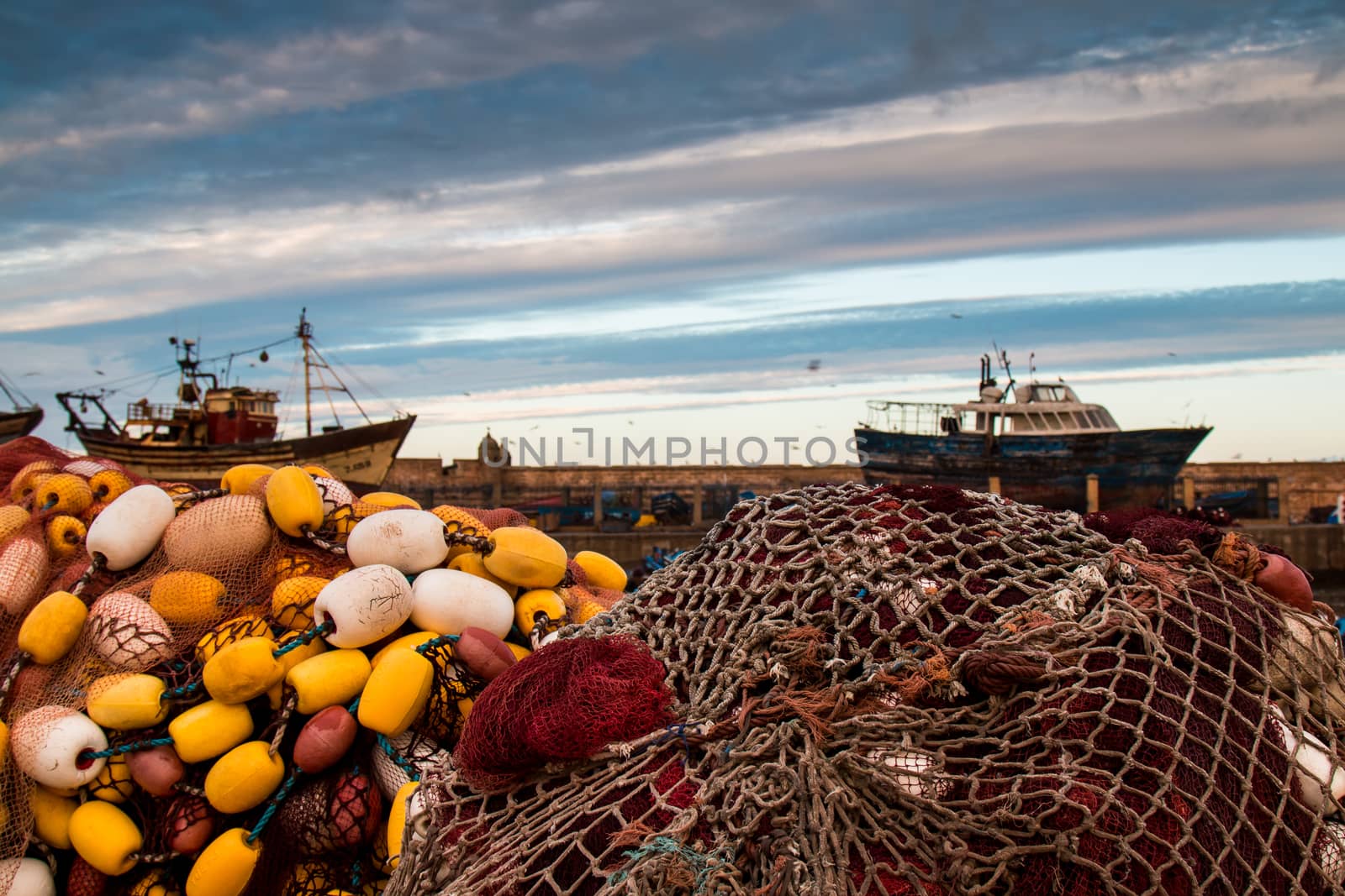 Early morning in the harbor. Pile of fishing nets, boats in the background. Dramatic sky with white and blue stripes. Essaouira, Morocco.