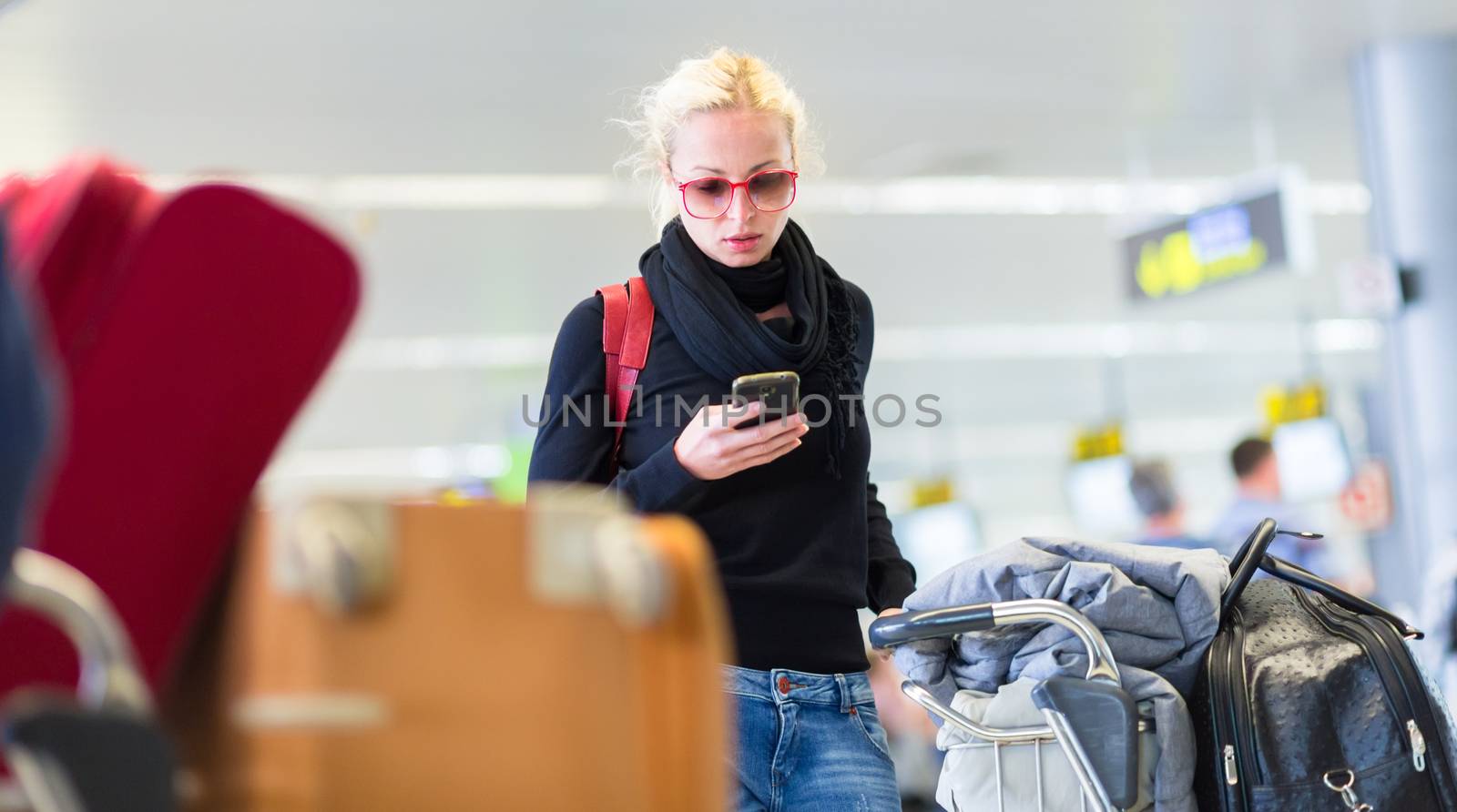 Casual blond young woman using her cell phone while queuing for flight check-in and baggage drop. Wireless network hotspot enabling people to access internet conection. Public transport.