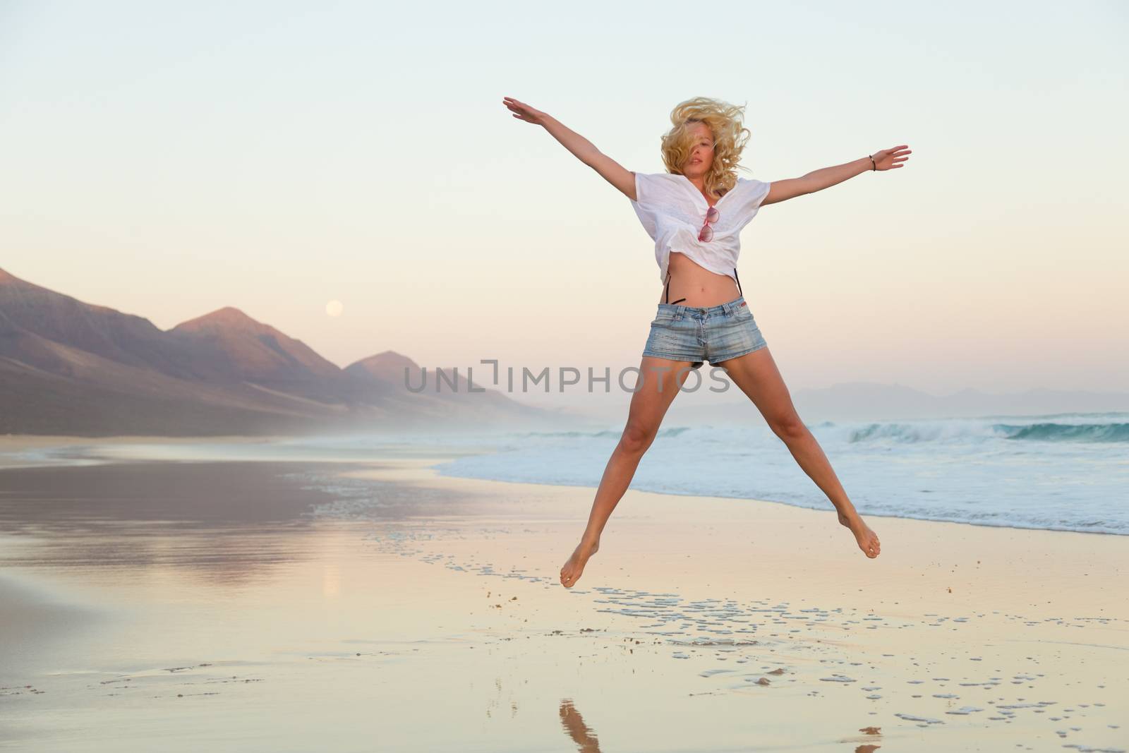 Young beautiful woman wearing jeans shorts and white t-shirt jumping on Cofete beach, Fuerteventura, Spain in sunset.