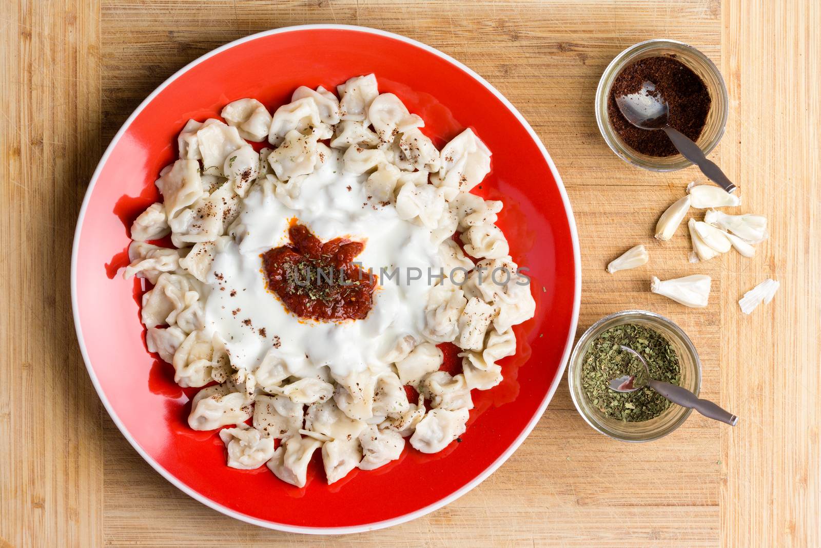 Delicious Turkish dumplings prepared and served on red plate next to seasonings in cups and chopped raw garlic