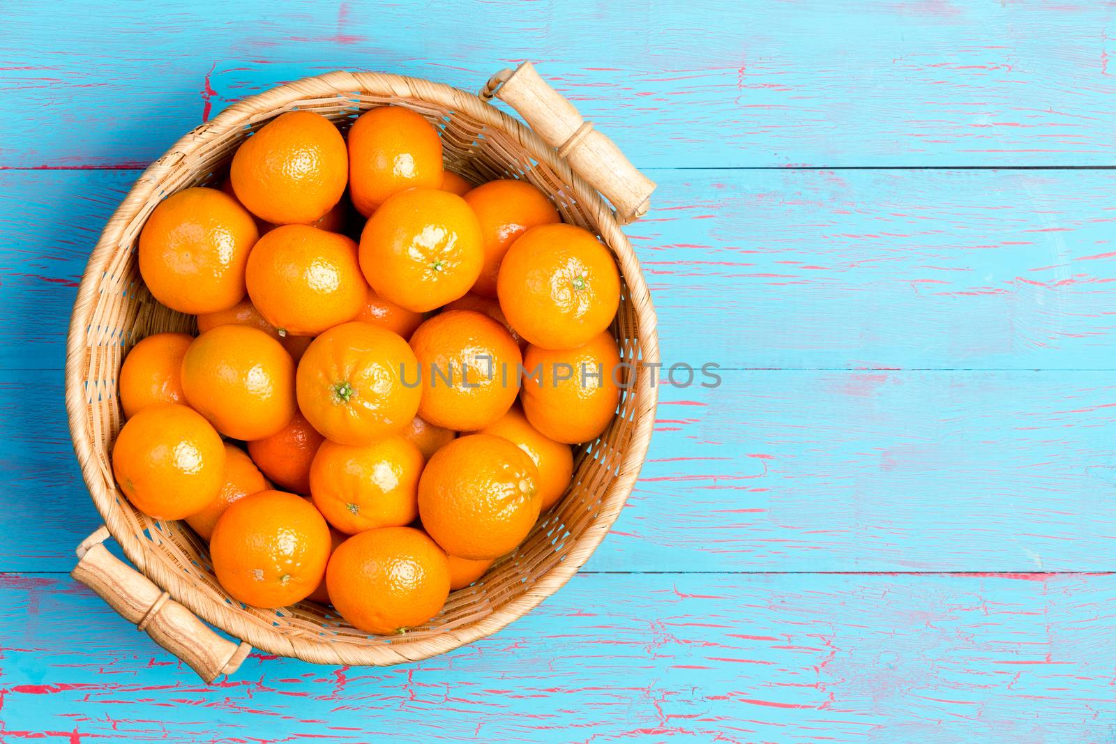 Top down first person perspective view of straw basket filled with clementine oranges over painted blue wooden table with copy space