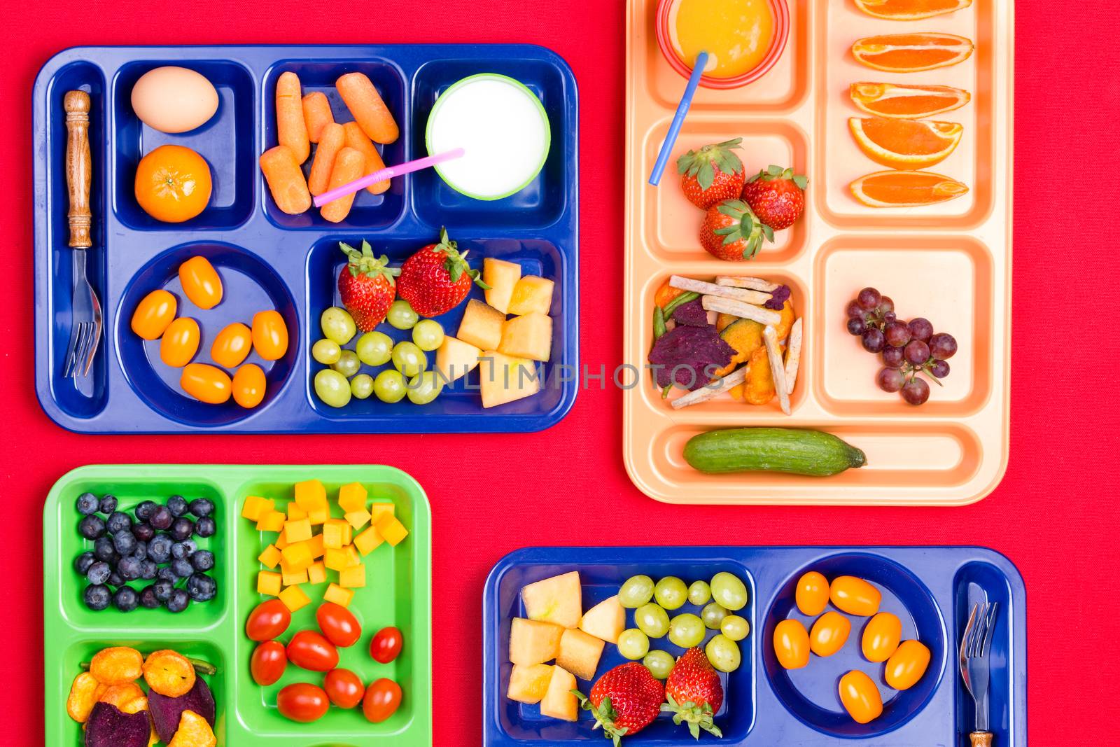 Four blue, green and pink plastic lunch trays filled with colorful fruit and vegetables along side milk and juice