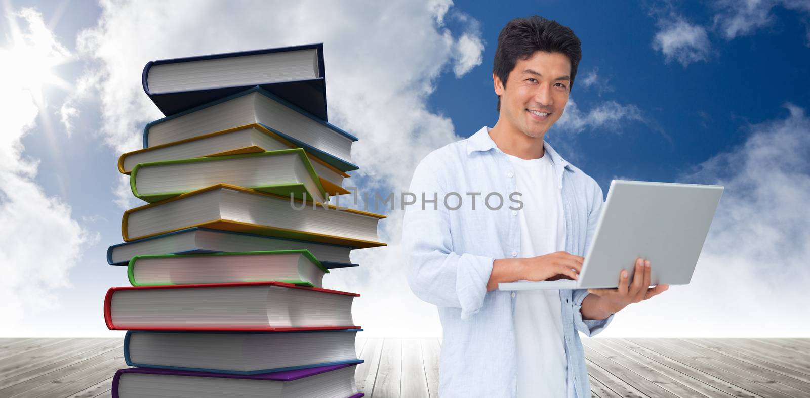 Smiling male with his laptop against stack of books against sky