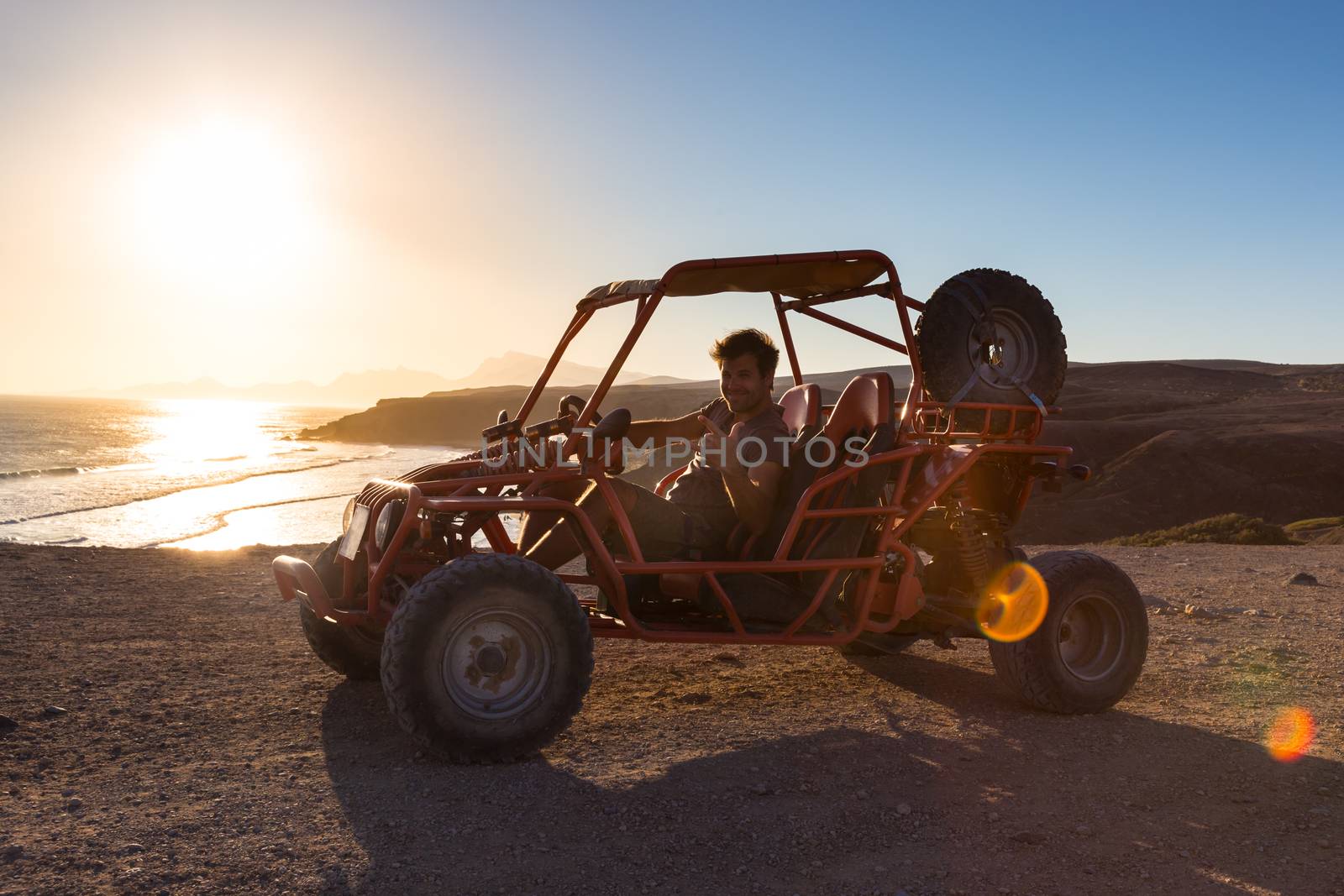 Active man driving quadbike on dirt road by the sea in sunset showing rocking sign to the camera.