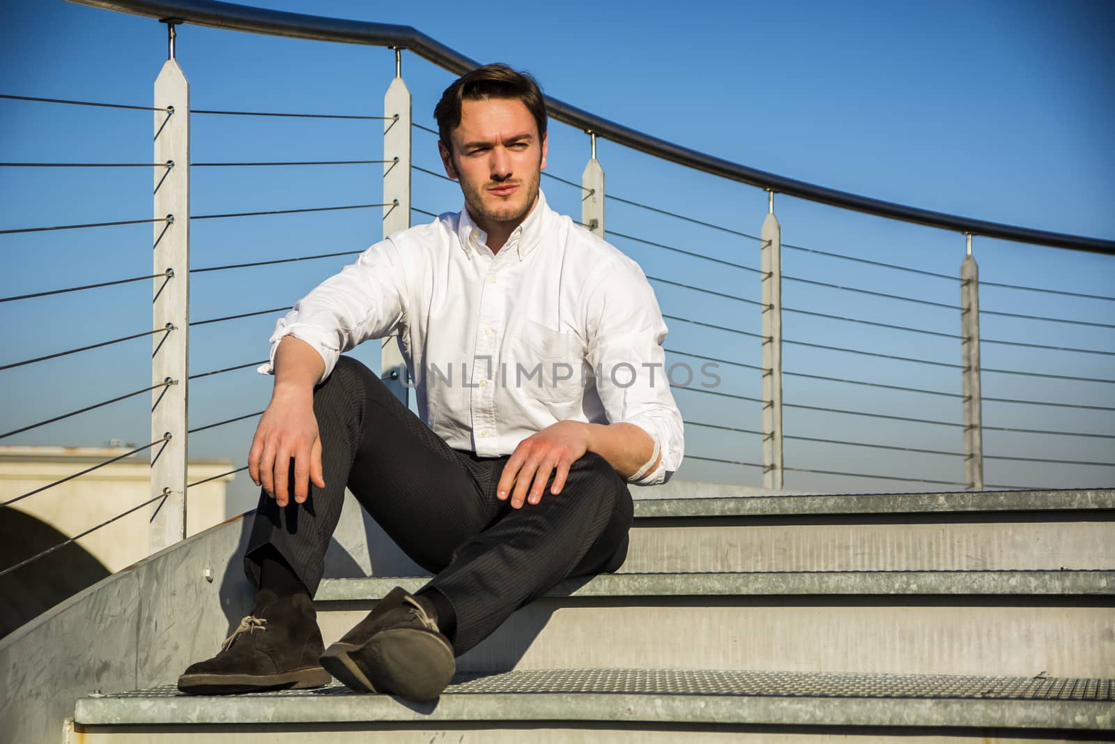 Handsome trendy man wearing white shirt sitting outdoor on metal stairs step, in city setting in a clear day