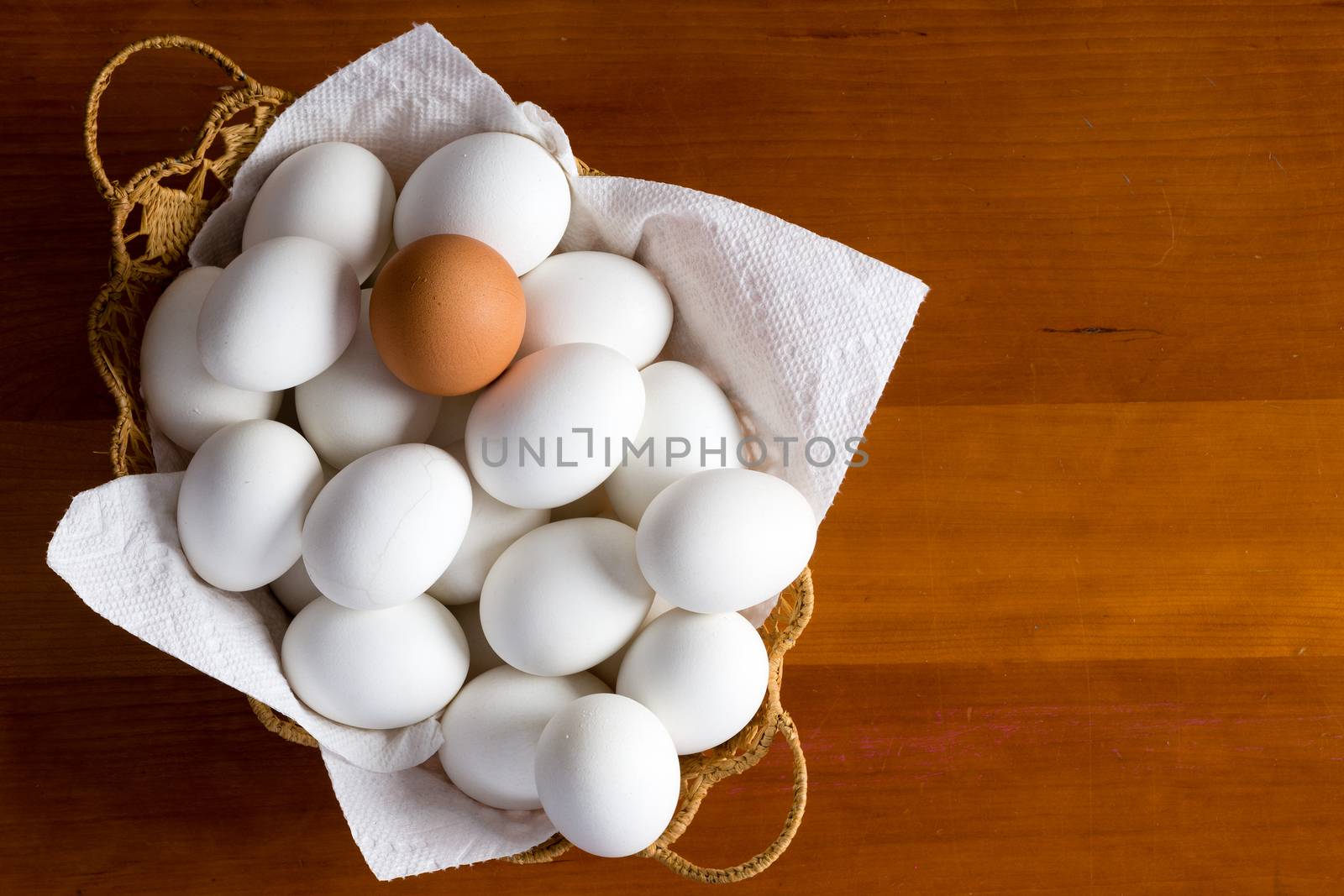 Wicker basket full of white eggs and single brown type on top over wooden table background with copy space on side