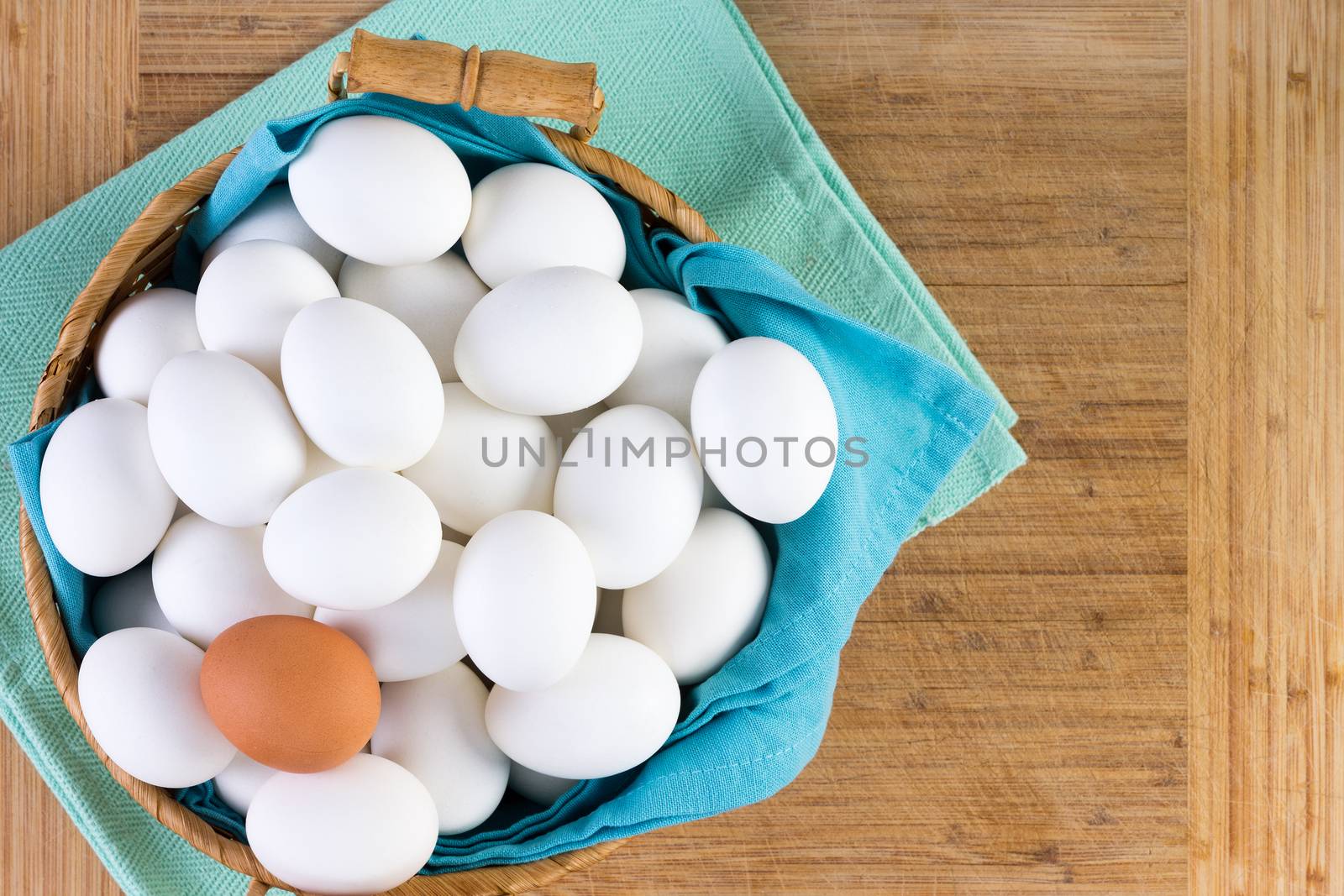 Wicker basket full of white eggs with one brown egg on the top in a conceptual image of diversity and individualism, overhead view on a wooden board background with copy space