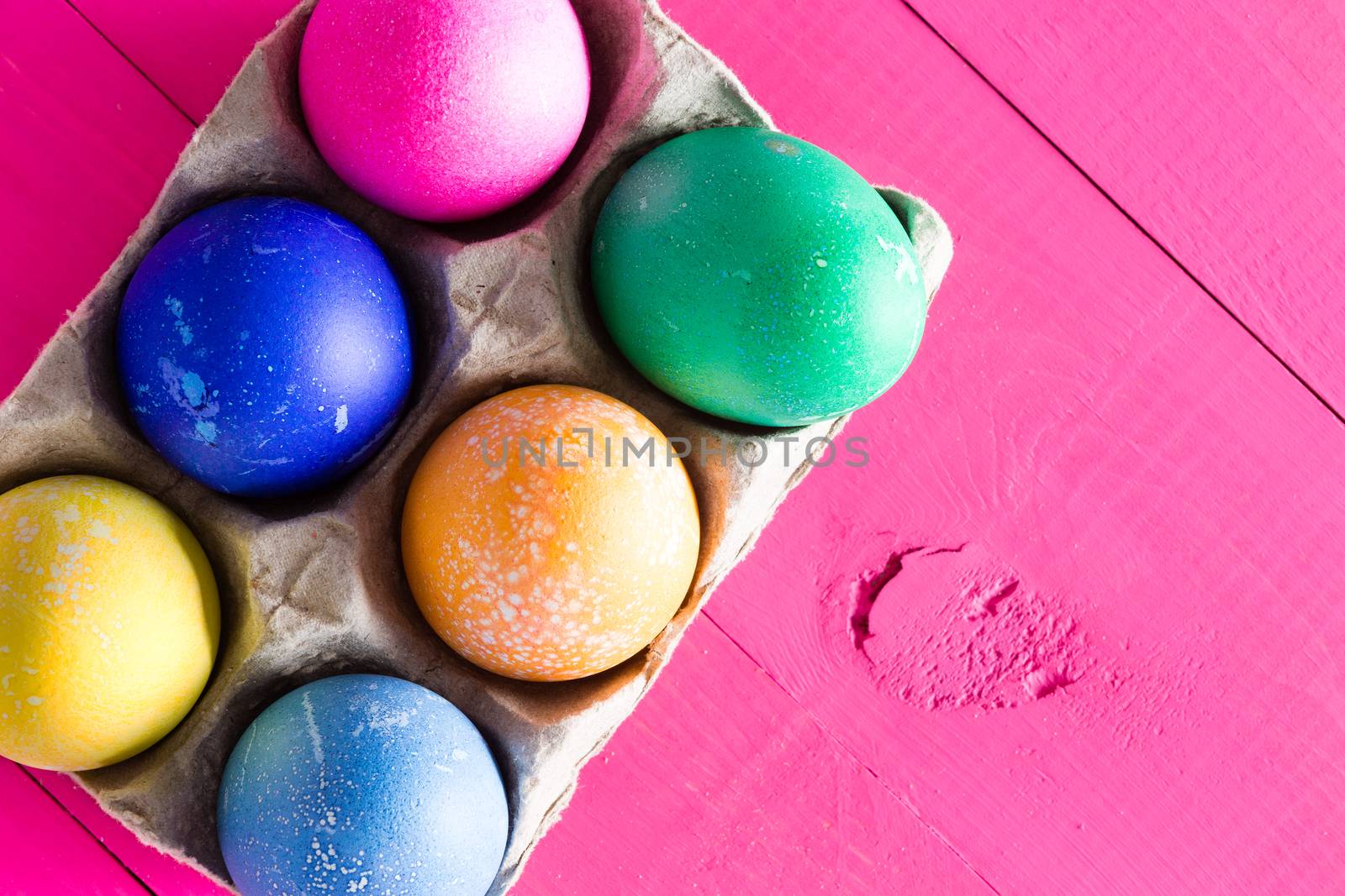 Six colorful hand decorated Easter eggs dyed with different colored pigments and displayed in a cardboard carton on an exotic magenta wooden background with copy space