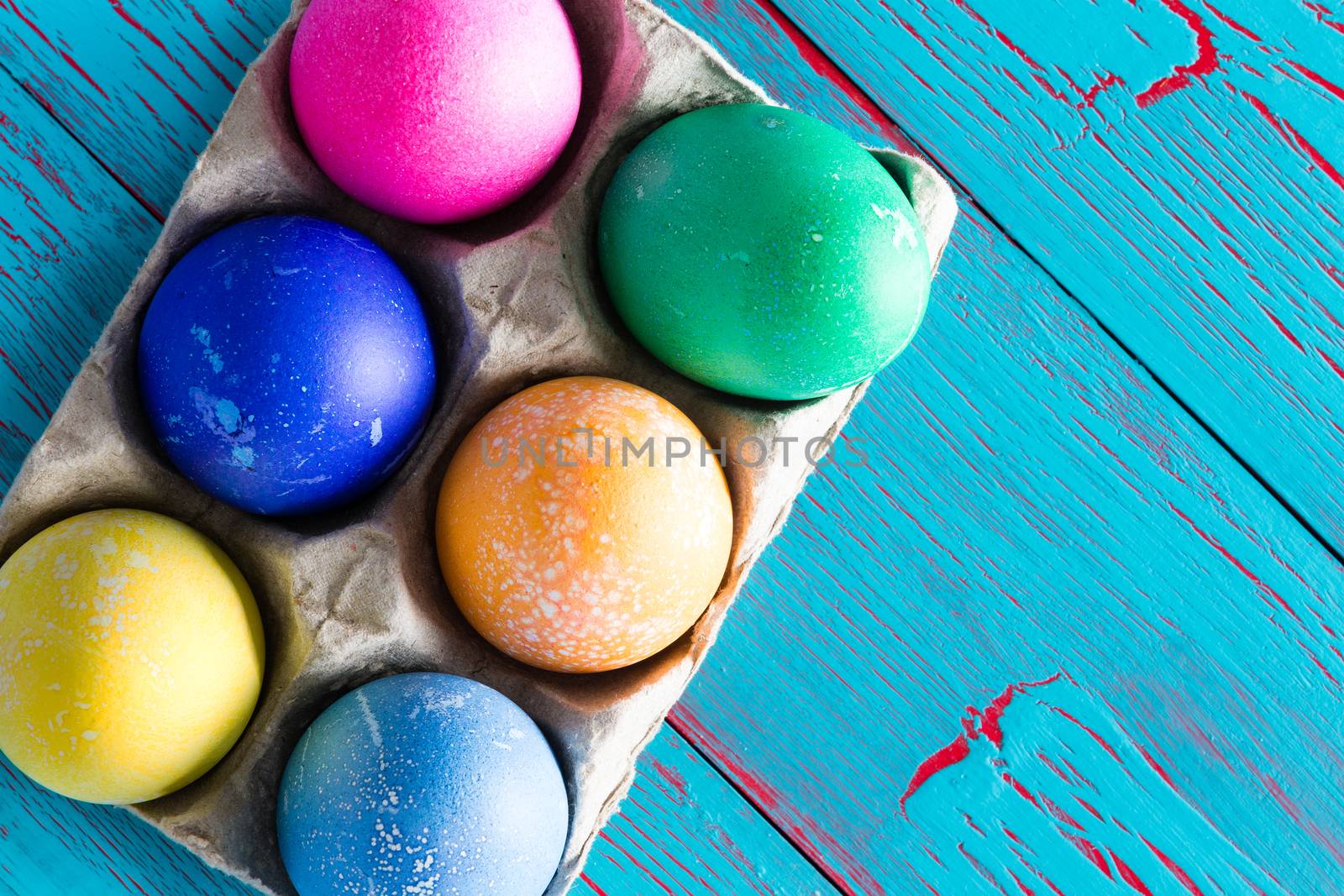 Six colored dyed Easter eggs in a cardboard egg box with copy space over a rustic exotic tropical blue wooden background with crackle paint, overhead view